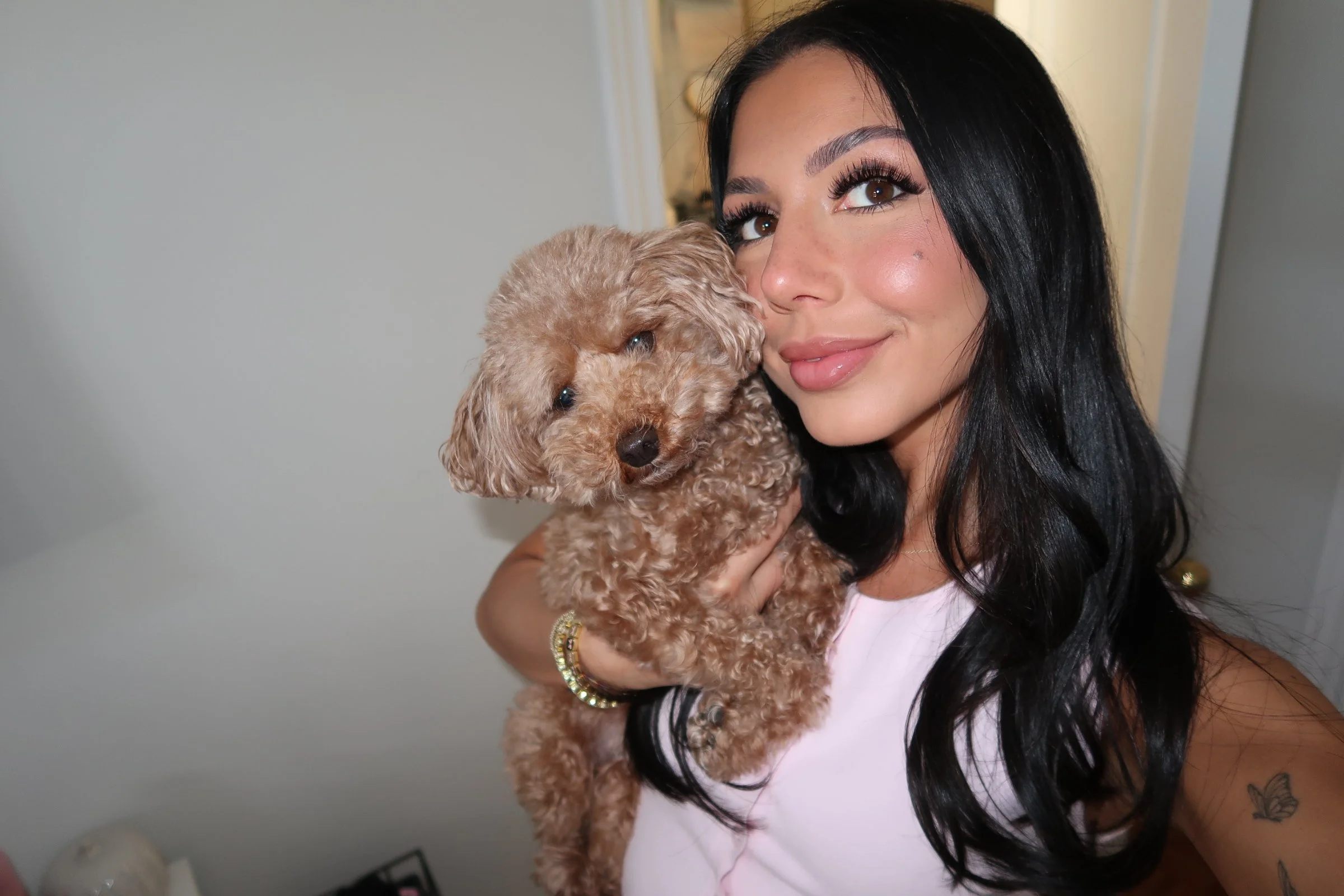 A woman with long black hair smiling while holding a small, curly-haired tan dog close to her face in a room with light-colored walls.