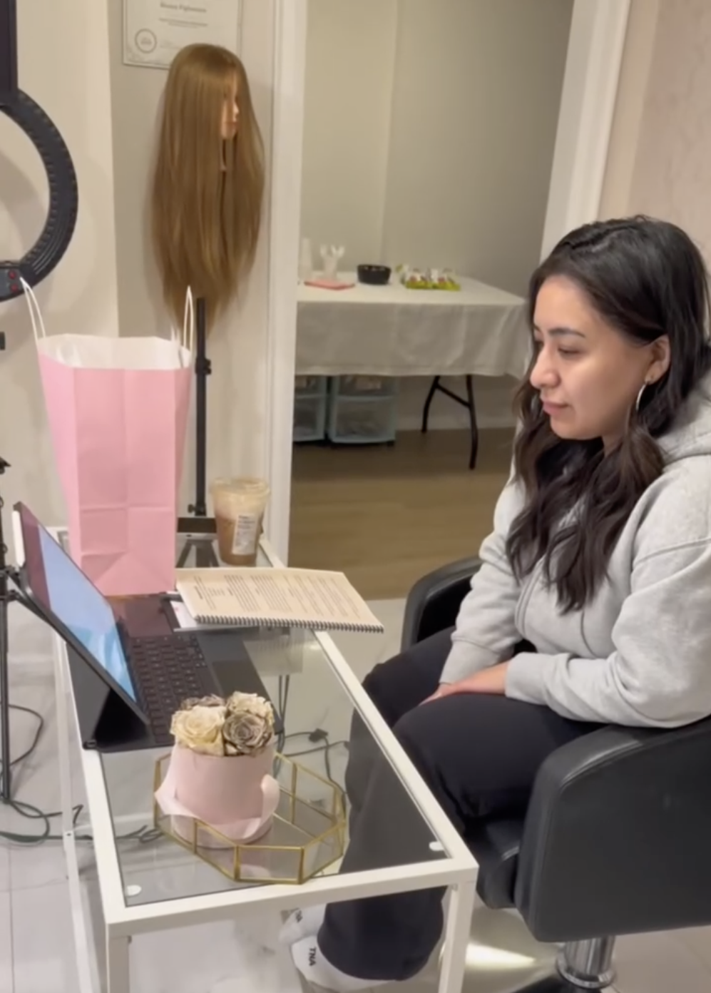 A woman sitting at a desk in front of a laptop and tablet, looking at the screen with a neutral expression, in a cozy home office or salon environment. On the desk are a pink flower arrangement, a pink gift bag, an open notebook, and a cup of iced coffee. A wig or mannequin head with long, reddish-brown hair is visible in the background.