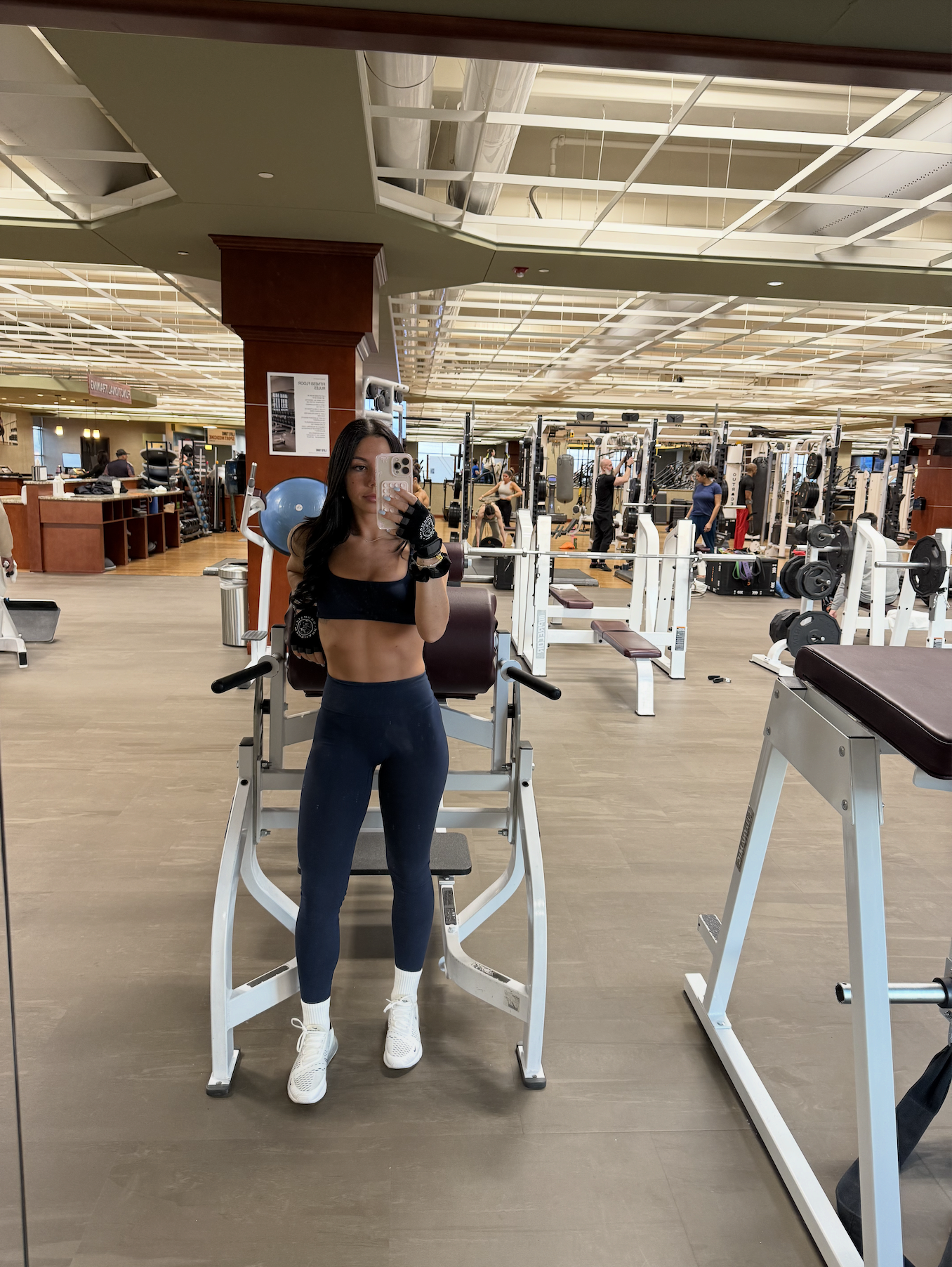 A woman taking a mirror selfie in a gym, wearing a black workout top and leggings. The gym has various exercise equipment and multiple people working out in the background.