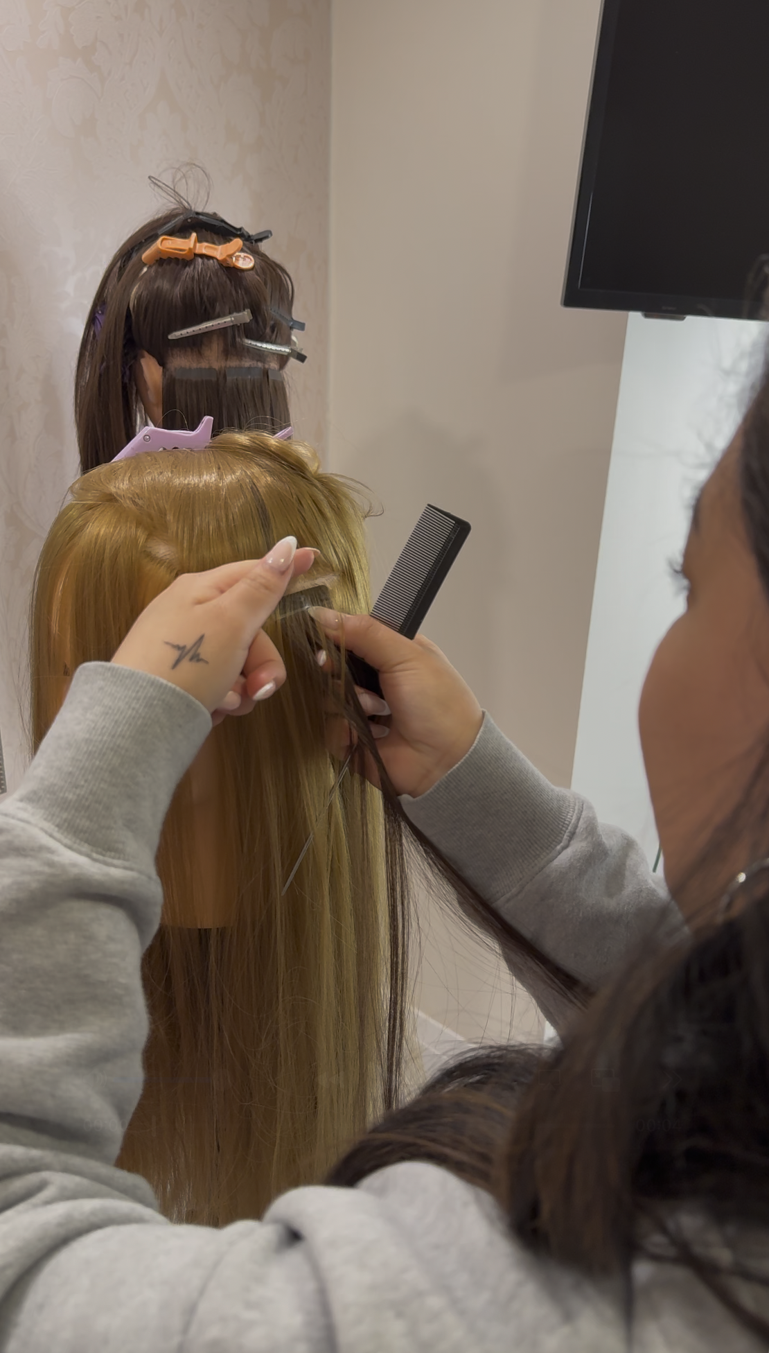 A hairstylist is cutting a client's long blonde hair while holding a comb and scissors. The client’s hair is sectioned, and hair clips are used to hold the sections. The background shows a mannequin head with styled hair and clips.