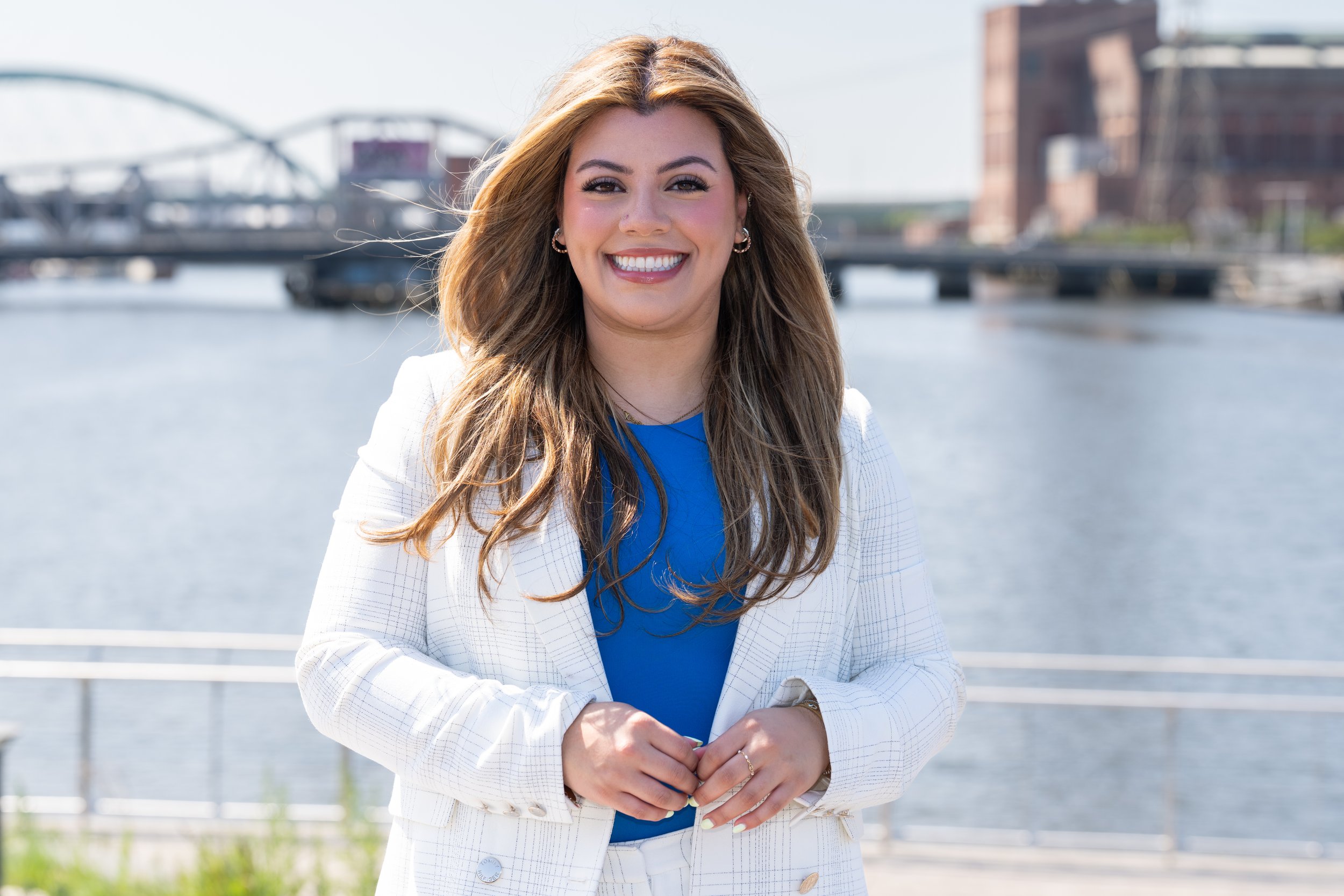A woman with long, wavy brown hair smiling outdoors near a river with the Providence skyline in the background, wearing a white blazer and a blue top.