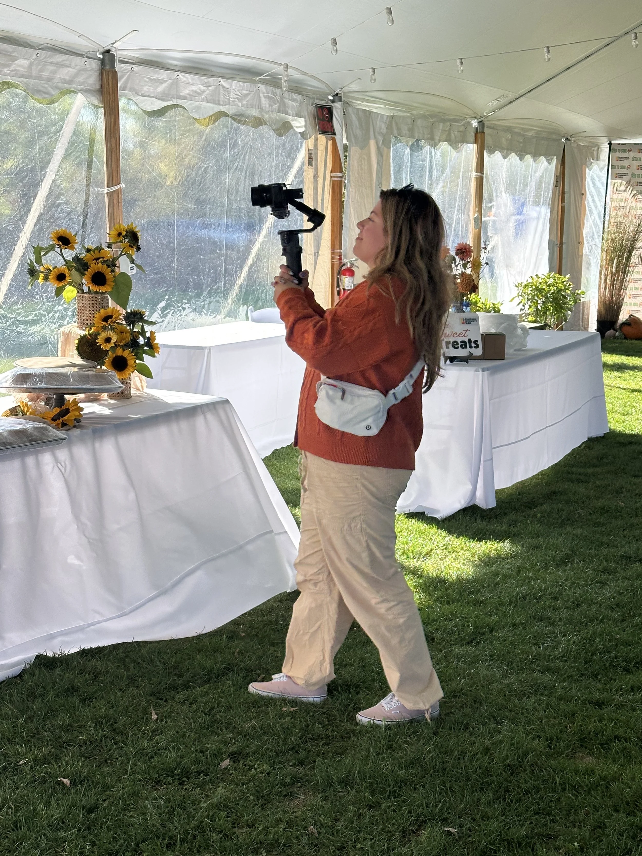 A woman in tan pants and an orange sweater is taking photos with a camera mounted on a stabilizer inside a white event tent decorated with sunflowers and other flowers on tables.