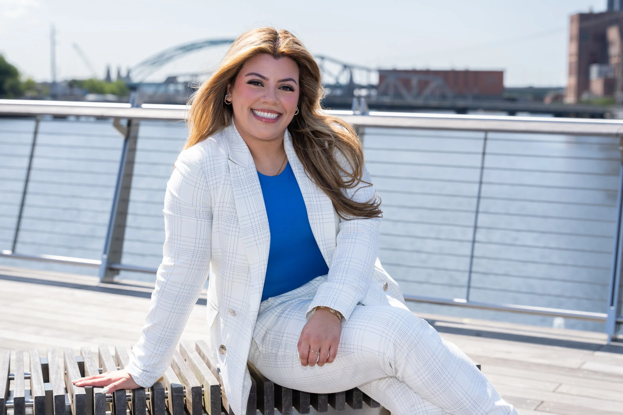 A woman with long, wavy hair in a white plaid suit and blue top sitting on a bench by a river, smiling at the camera.