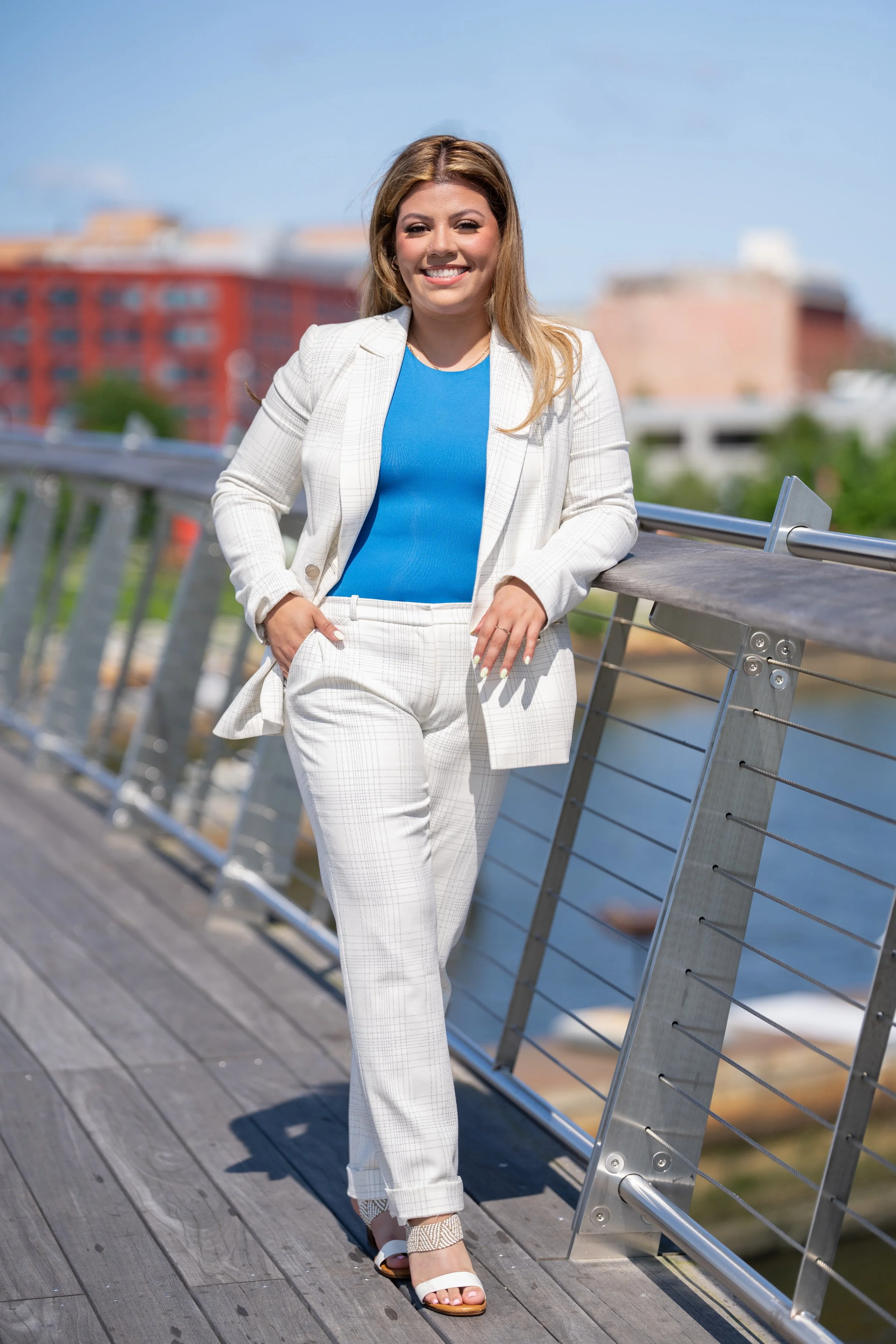 A woman in a white plaid suit and blue top leaning on a metal railing outdoors on a sunny day, with the Provience cityscape and water in the background.