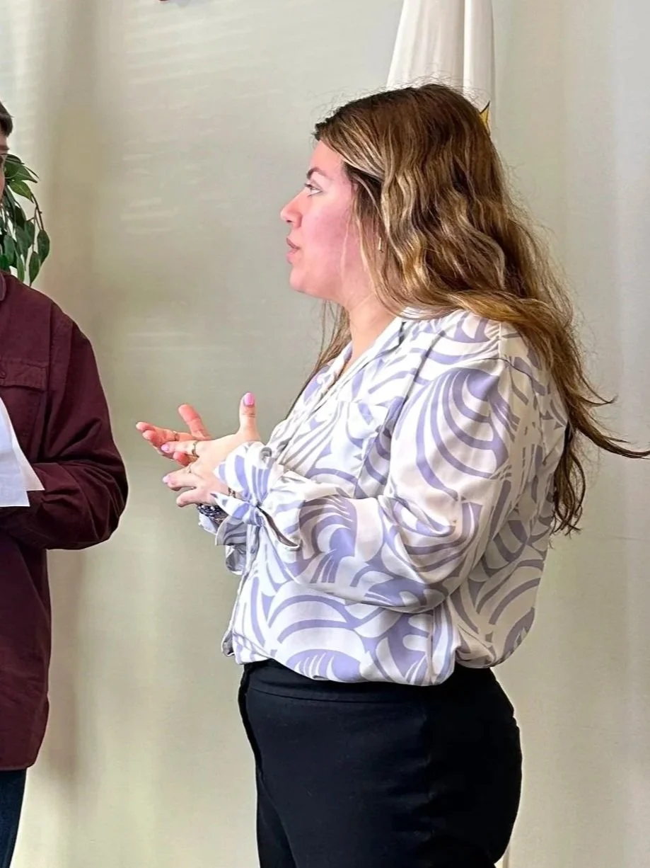 A man and a woman are having a conversation in an indoor setting. The man is holding papers and looking at the woman, who is gesturing with her hands. There is an American flag, a fire alarm, and indoor plants in the background.