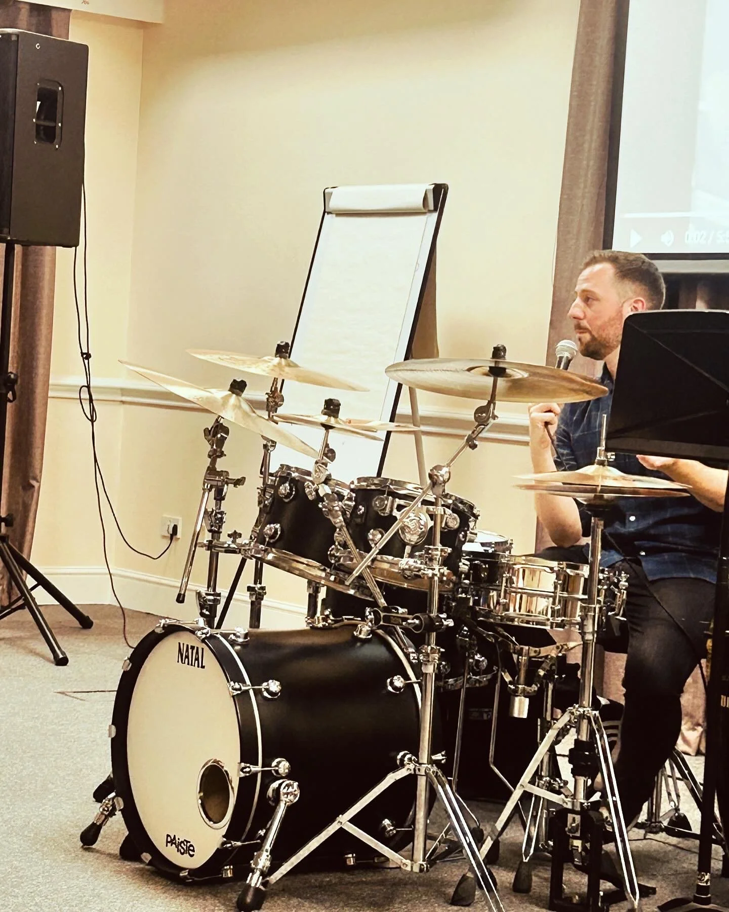 A man sitting behind a black drum set with cymbals, holding a microphone during a performance or rehearsal in an indoor setting.
