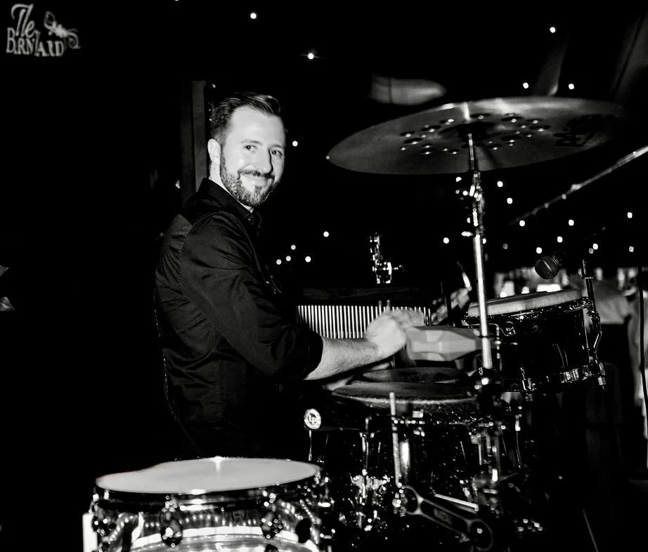 A smiling man playing a drum set in a dimly lit venue with small lights in the background.