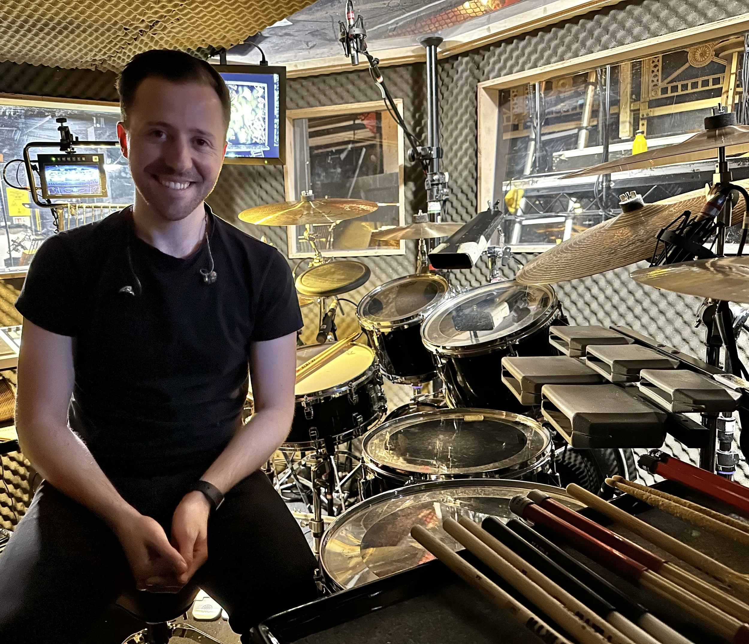 A smiling man sitting in a soundproofed music studio, surrounded by electronic drums and percussion instruments, with soundproof foam walls and recording equipment in the background.