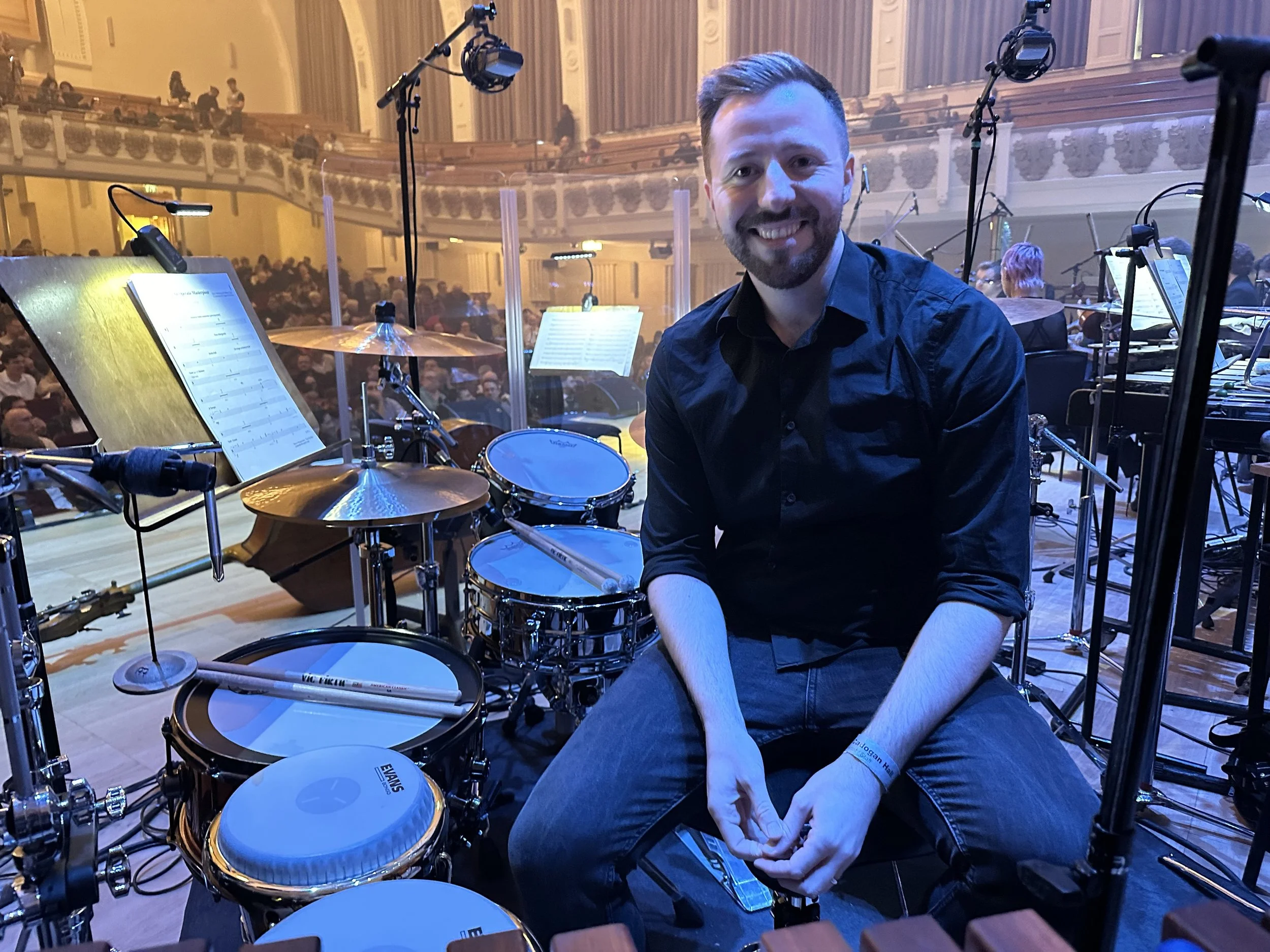 A young man sitting on the stage behind a drum set in a concert hall, smiling at the camera.