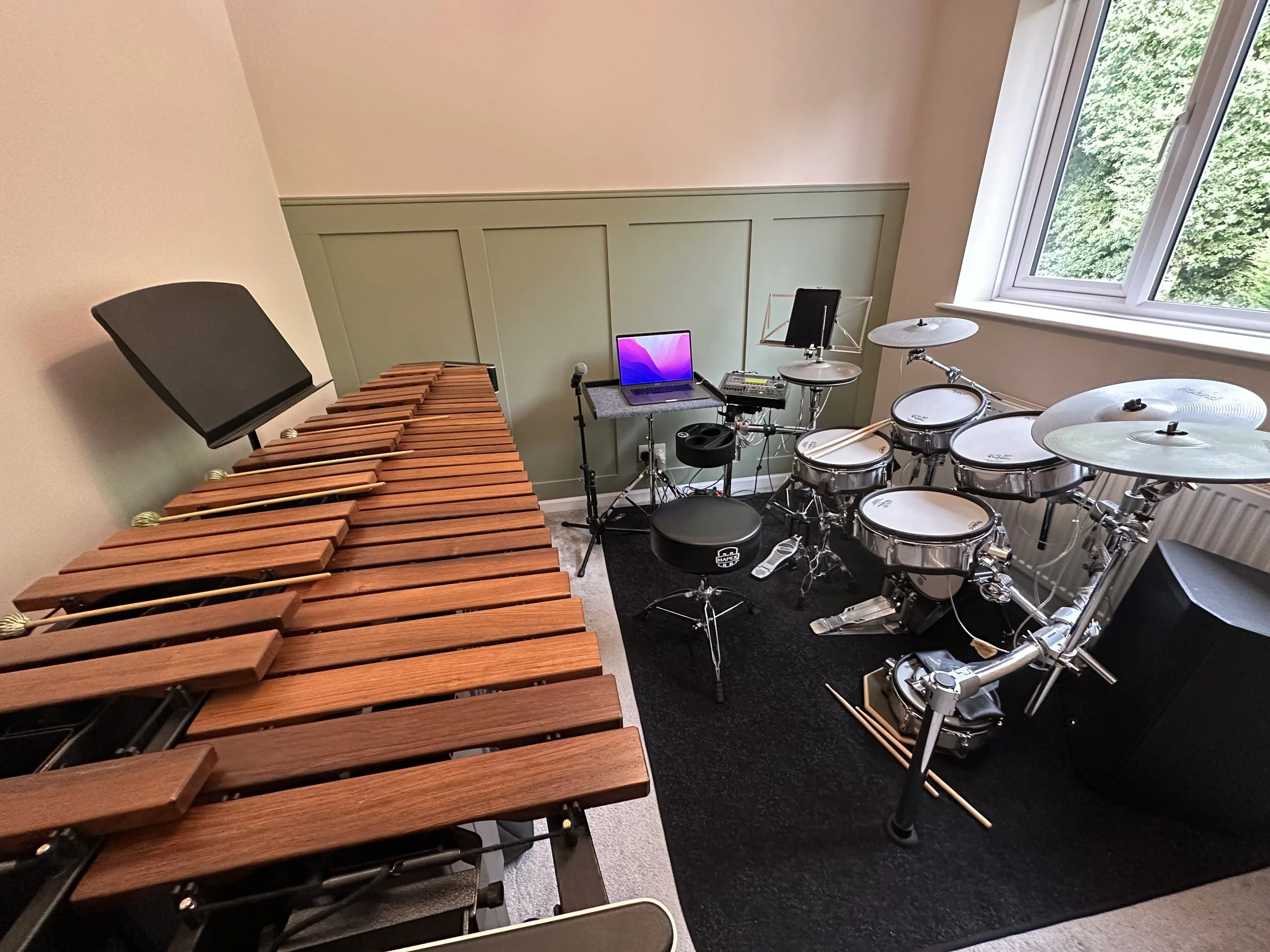 A music practice room with a digital drum set on a black rug, a marimba, a music stand, and a laptop on a small table near a window with a view of green trees.