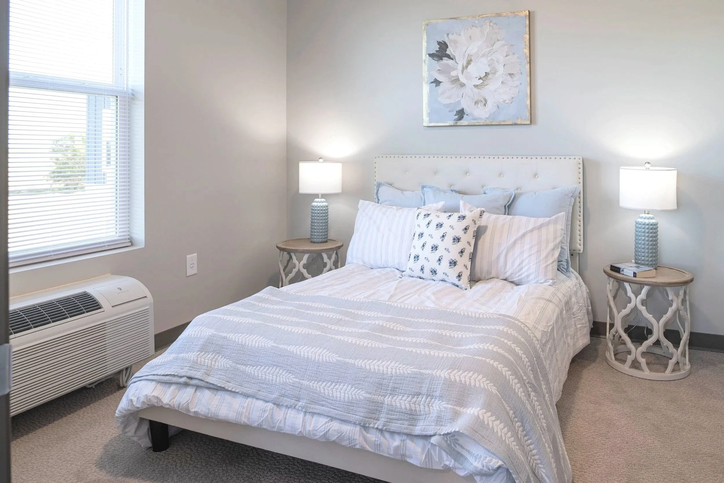 Bedroom with a white bed, light blue pillows, a framed floral painting above the bed, two matching nightstands with lamps, a window with blinds, and an air conditioner below the window.