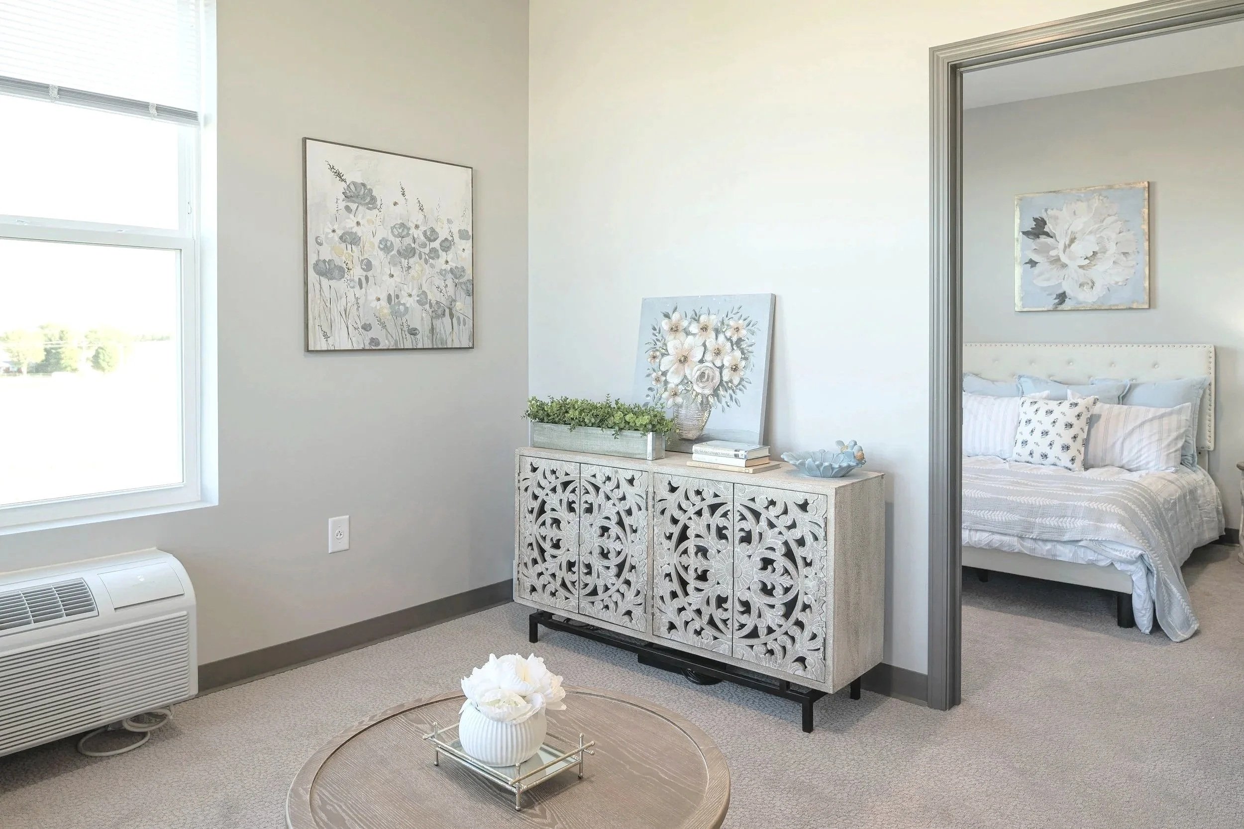 A bright bedroom with light-colored walls and decor, a bed with white bedding and pillows, a painting of flowers on the wall, a decorative white cabinet with floral carvings, and a small round table with a white flower arrangement.