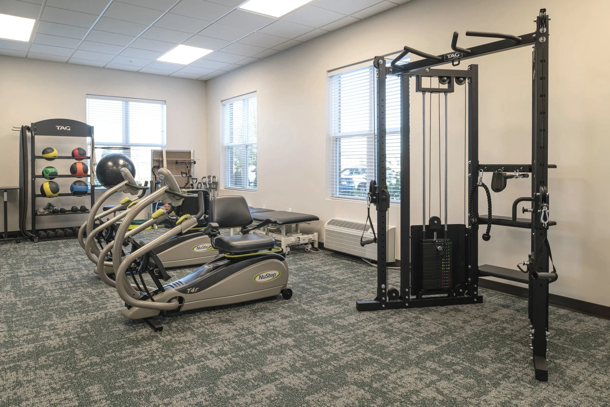 A small fitness room with exercise machines, weights, a stability ball, a rack of medicine balls, and a multi-purpose workout station.