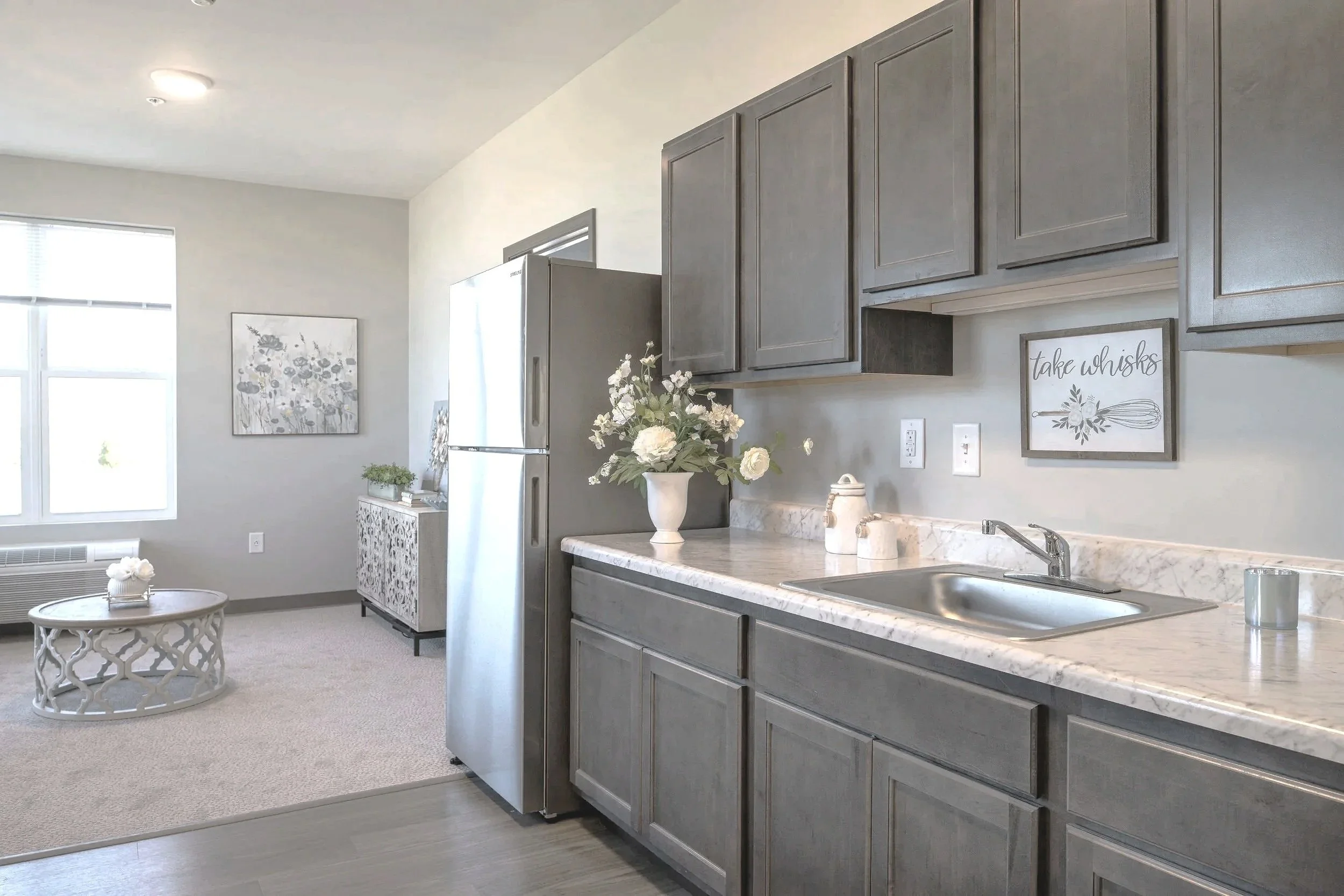Modern kitchen with gray cabinets, marble countertop, stainless steel sink, white flowers in a vase, and decorative signs on the wall, adjacent to a living area with a round coffee table and artwork on the wall.