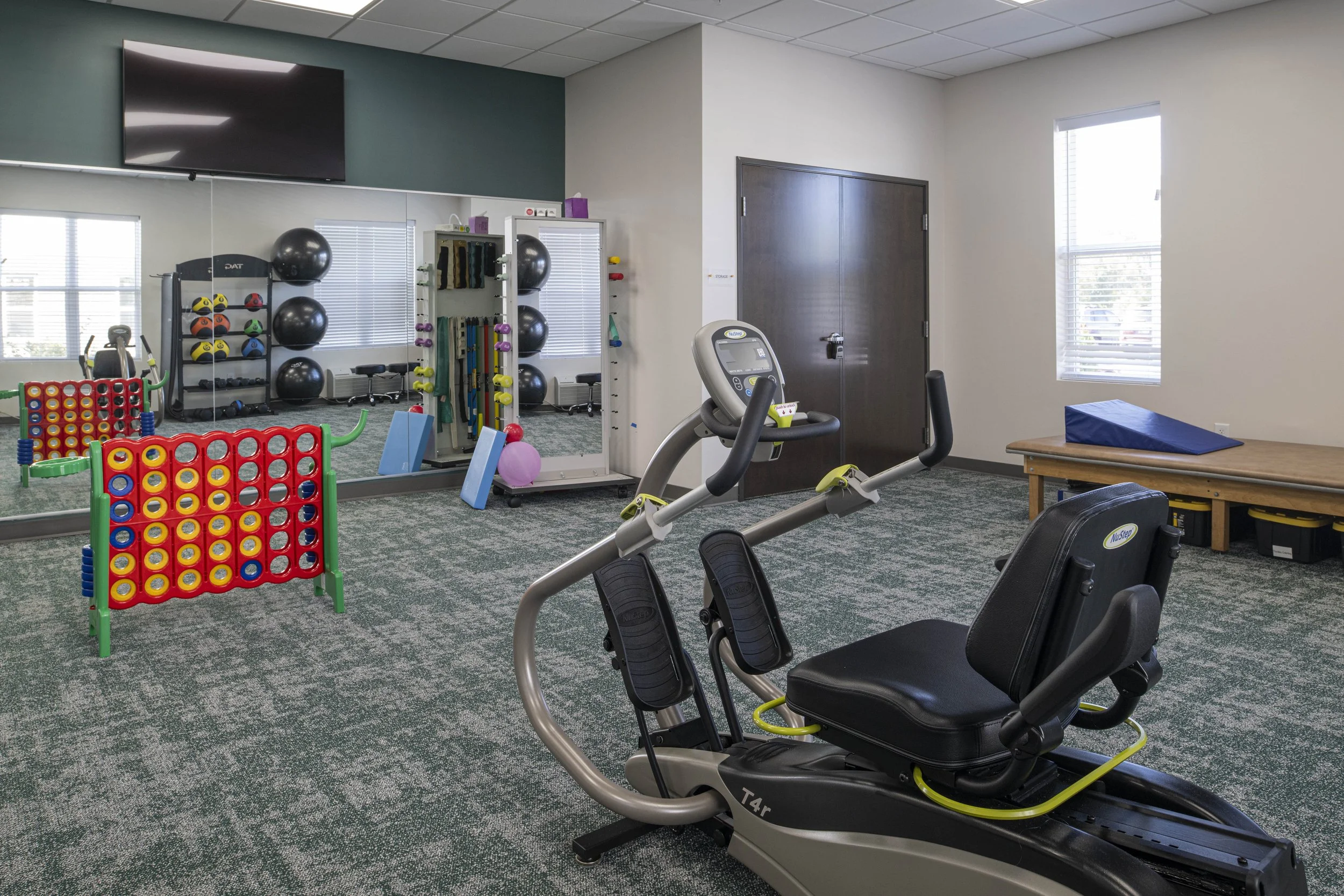 Interior of a physical therapy or rehabilitation gym with exercise equipment, including a recumbent exercise bike in the foreground, a large mirror on the wall, and exercise accessories like balls, weights, and foam pads arranged around the room.