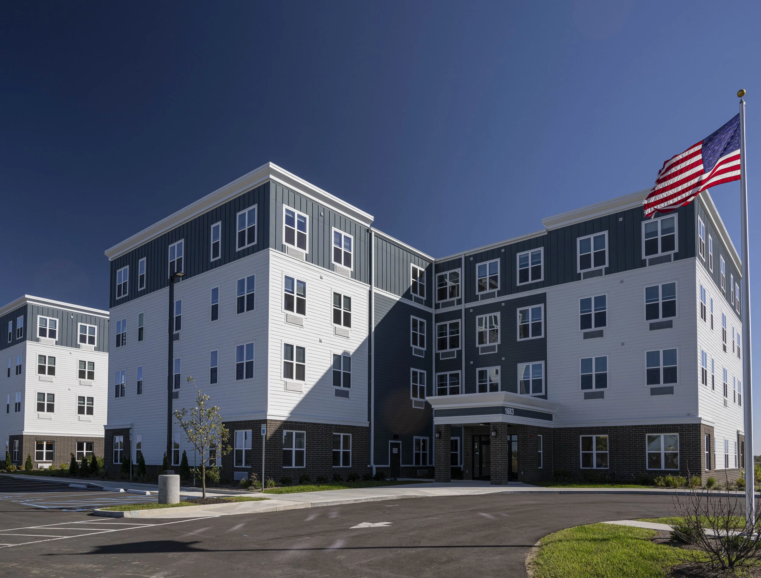 A modern multi-story apartment building with white and dark blue exterior, multiple windows, and an American flag on a flagpole in front of it.