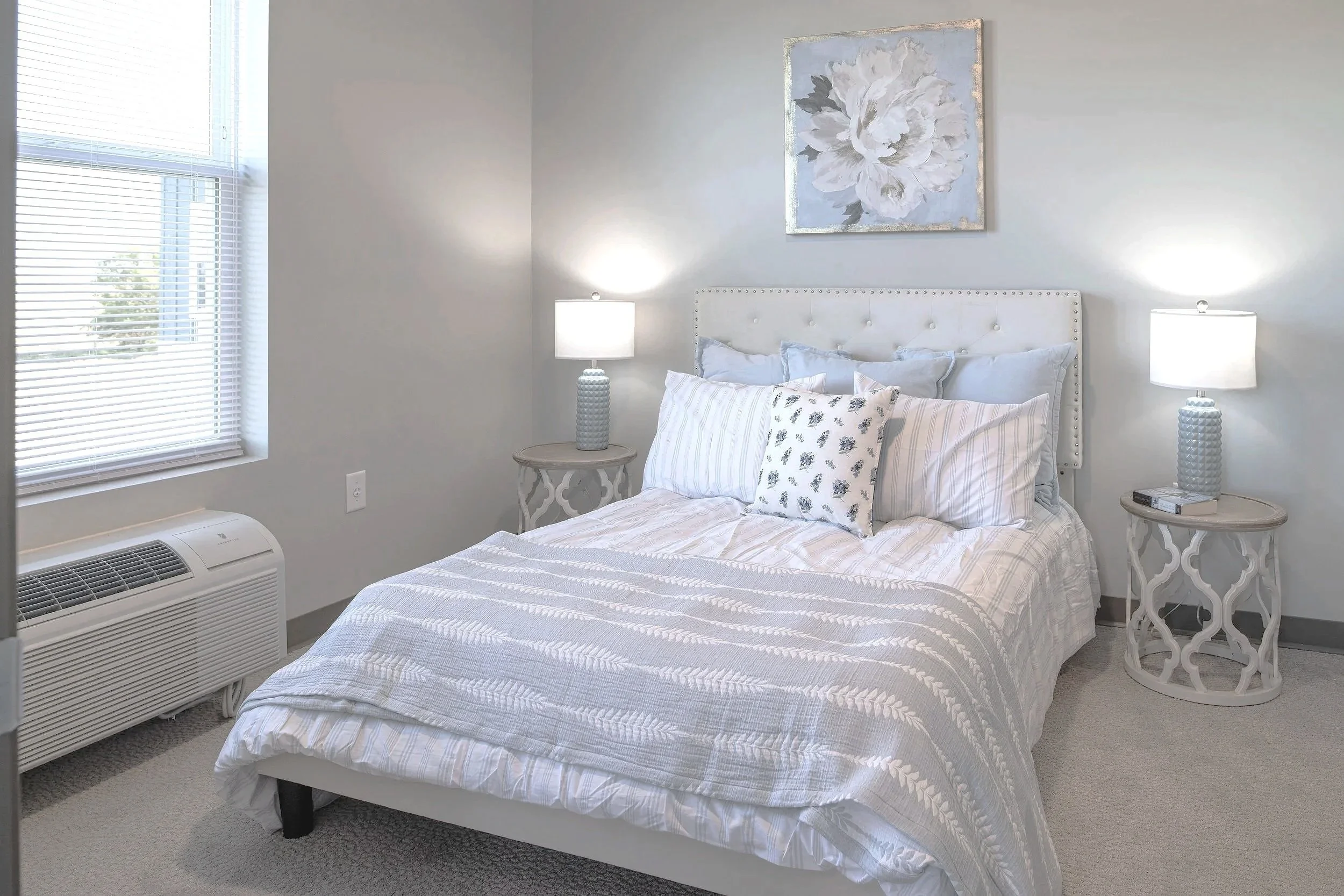 A neatly made bed with white and light-colored bedding, positioned against a wall with a large floral painting. There are two bedside tables with matching lamps, a window with blinds, and an air conditioning unit below the window.
