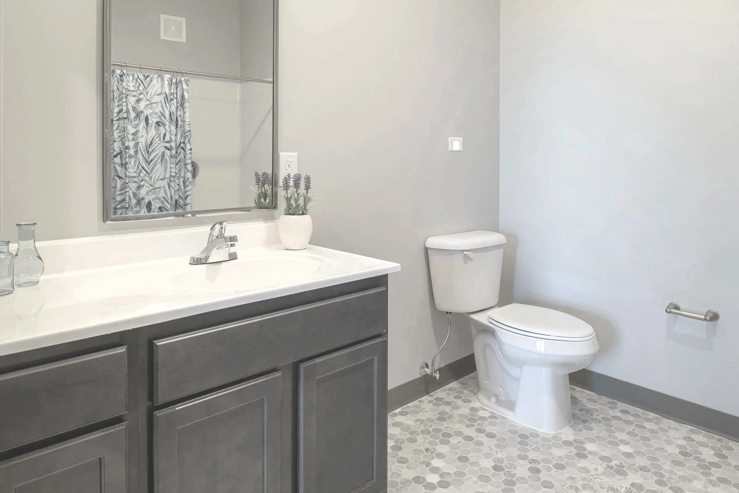 Bathroom with gray vanity, white countertop, large mirror, potted lavender plant, toilet, and hexagonal tile floor.