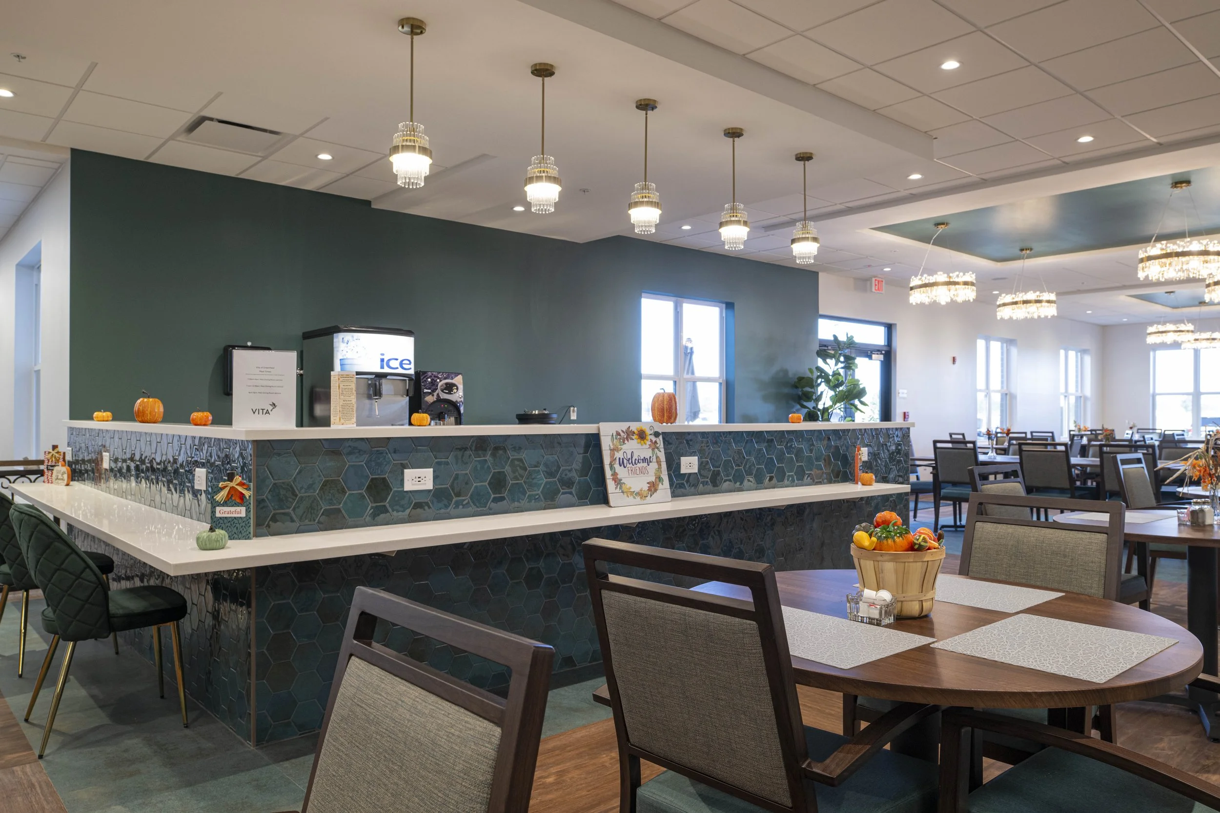 A restaurant dining area with wooden tables, beige and green chairs, and a decorated counter with pumpkins and a welcoming sign, illuminated by elegant chandeliers and natural light from windows.