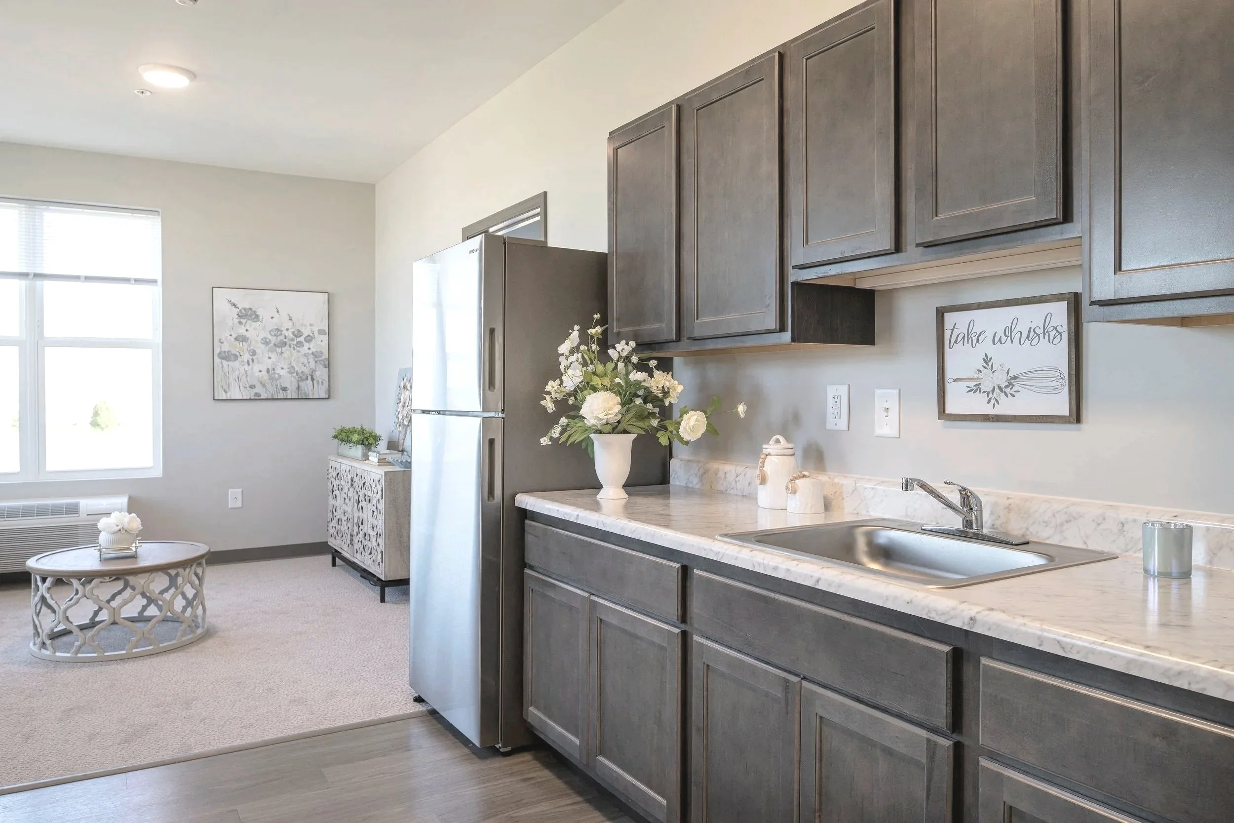 Modern kitchen with gray cabinets, marble countertops, a stainless steel sink, and decorative flower arrangements. An open living space with a window, artwork, and a round coffee table is visible in the background.