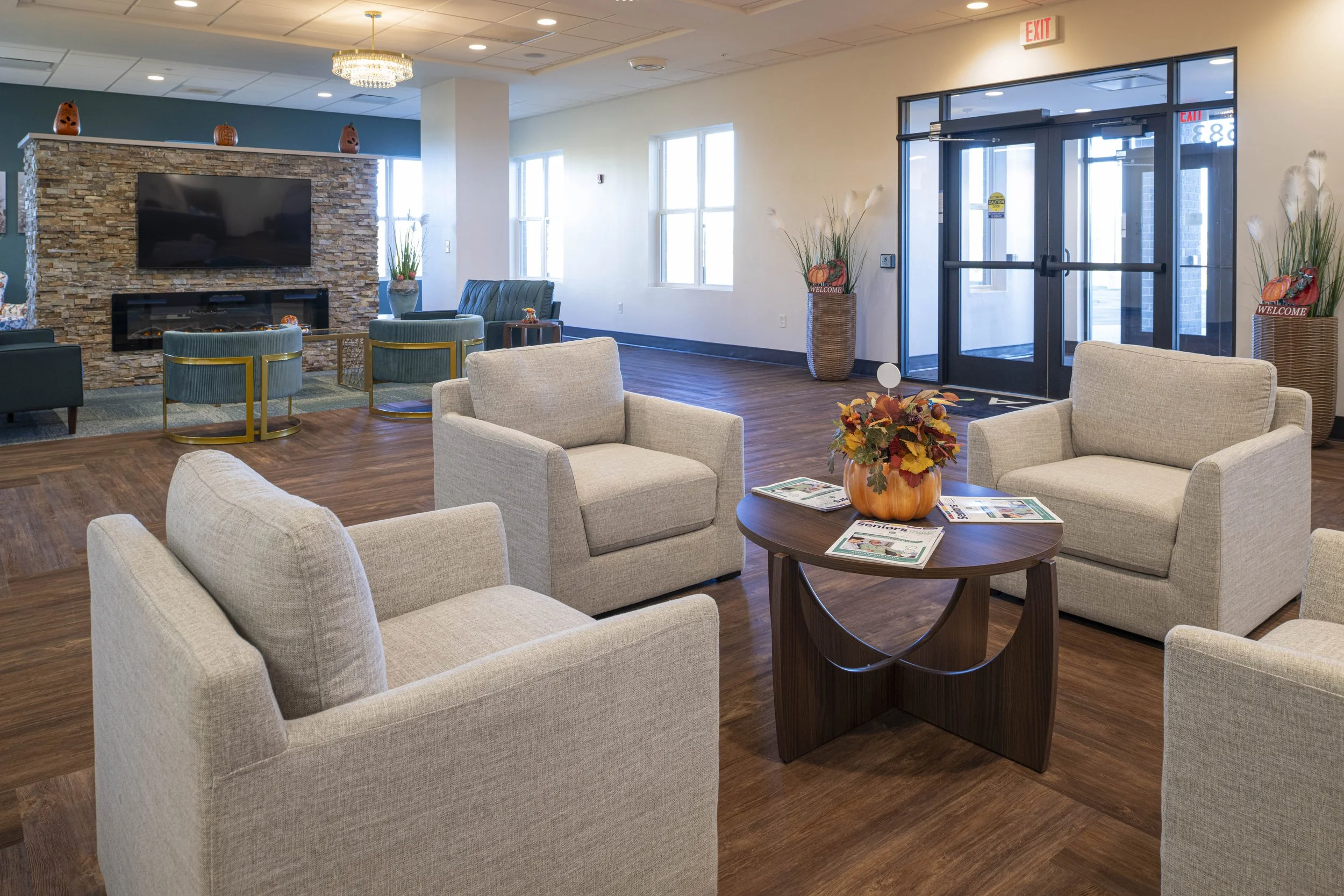 A cozy waiting area with four beige armchairs arranged around a round wooden table with autumn-themed decorations and magazines. In the background, there is a seating area with teal armchairs and a stone fireplace with a large TV above. The room features hardwood flooring, decorative plants in vases, and a glass entrance door with exit signage.