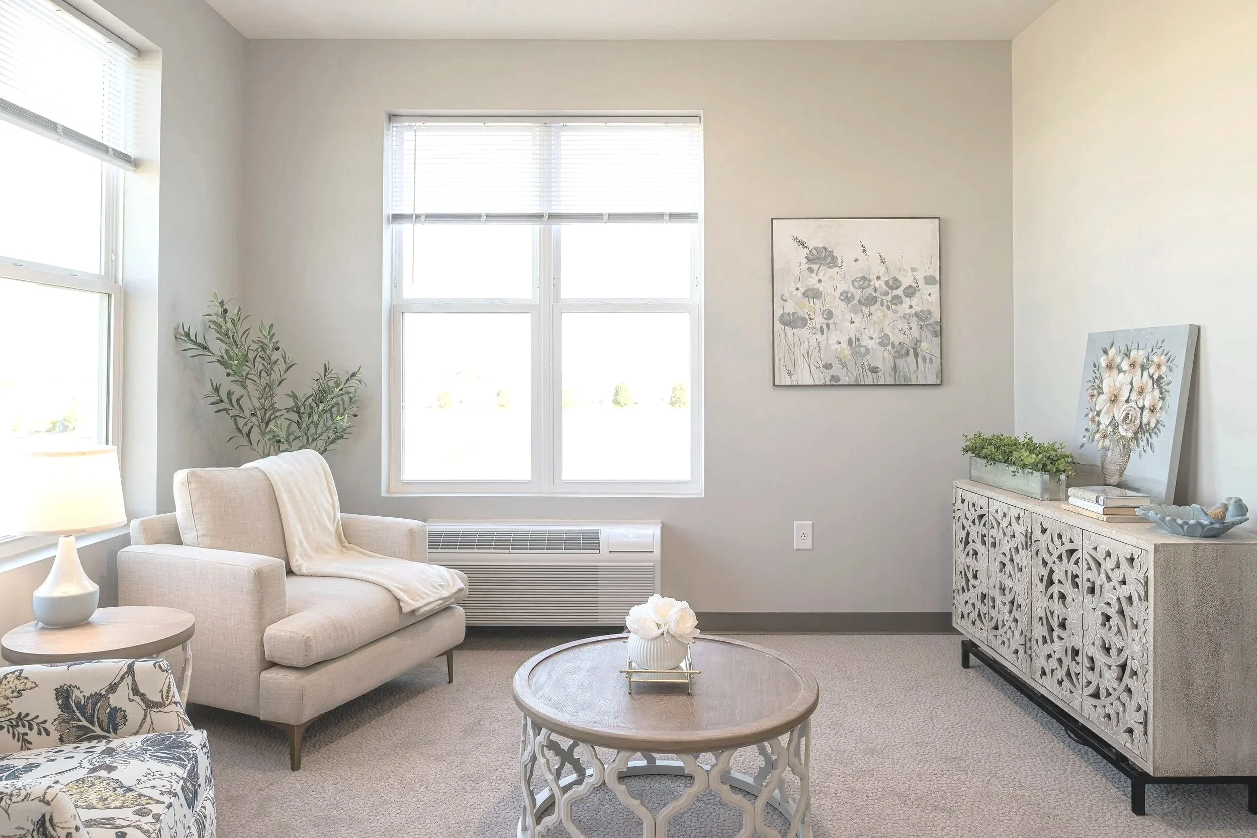 A bright living room with a beige sofa, a patterned armchair, a round wooden table with a white flower arrangement, a white ornate sideboard with decorative items, large windows, and framed floral art on the walls.
