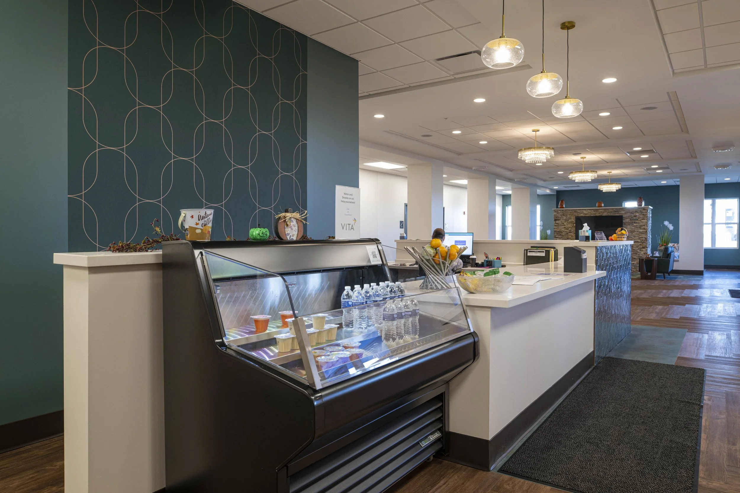 Lobby reception area with a white counter, ice cream freezer, and a computer station in a modern building with ceiling lights and a dark wood floor.