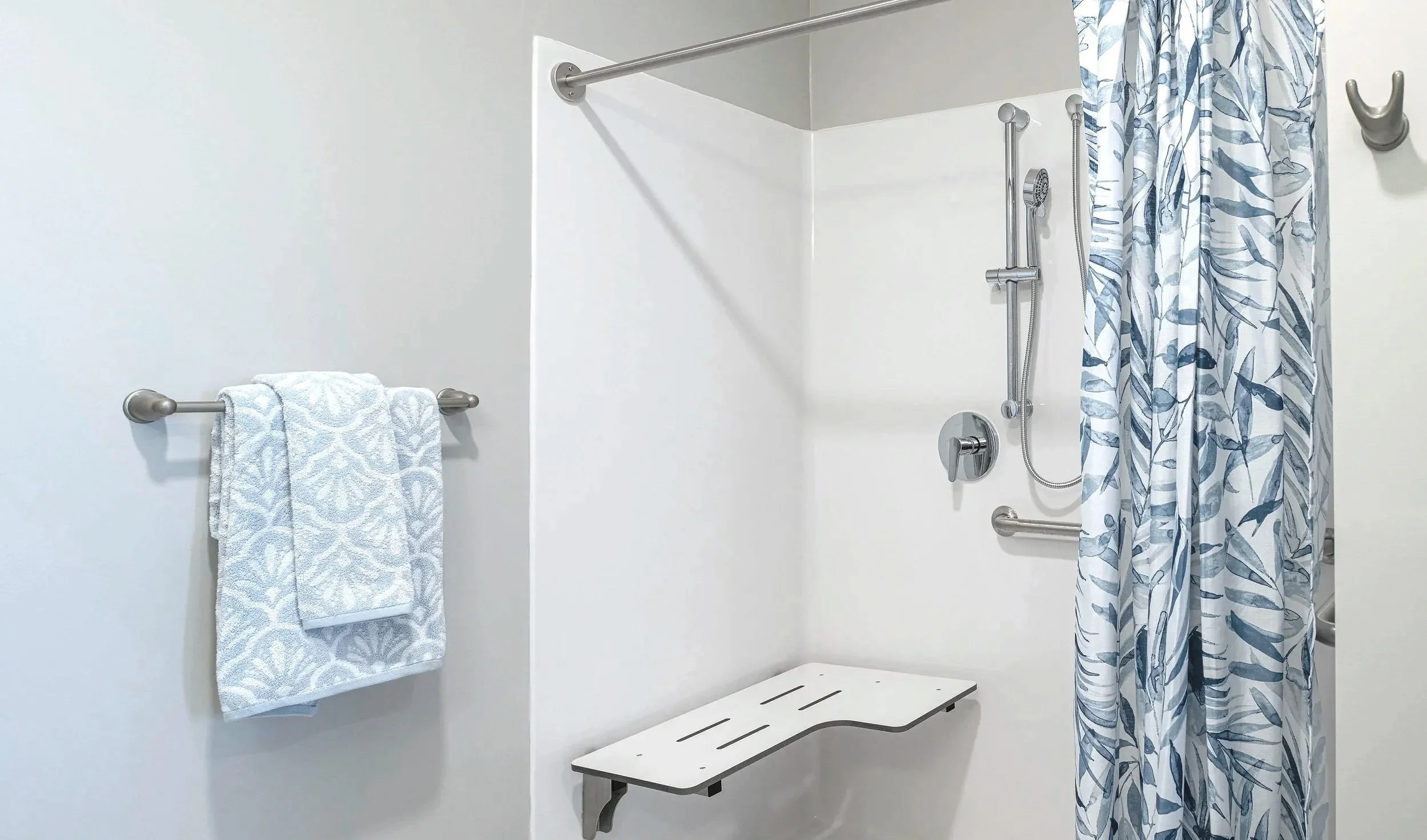 Accessible shower with a fold-down seat, grab bar, handheld showerhead, and a patterned shower curtain.