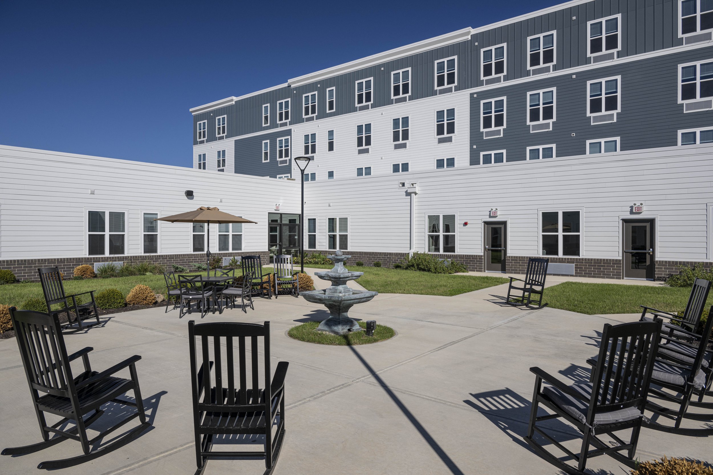 Outdoor courtyard with black rocking chairs, a fountain, and a patio table with an umbrella, surrounded by a modern multi-story building with white and gray siding