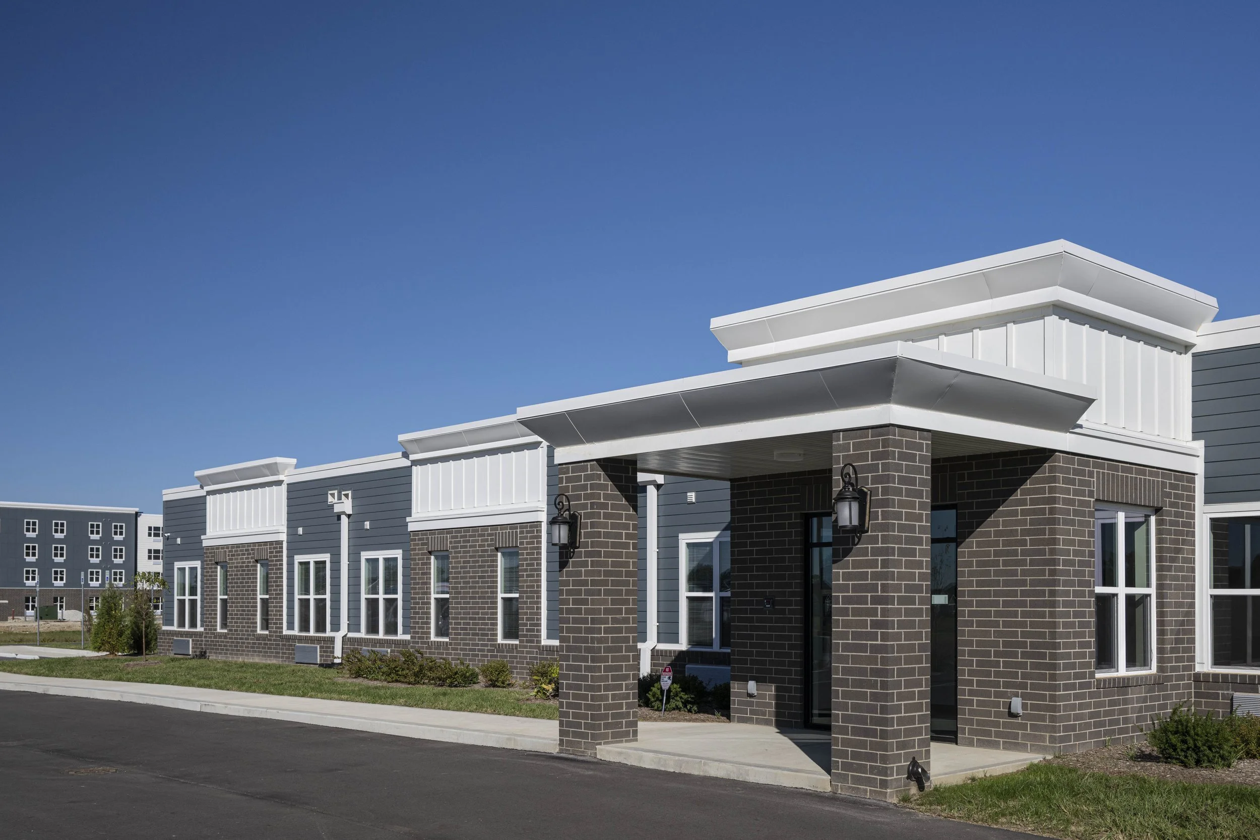 A modern apartment complex with gray siding, brick accents, and white trim, featuring multiple windows, entryway with overhead cover, and exterior lighting fixtures.
