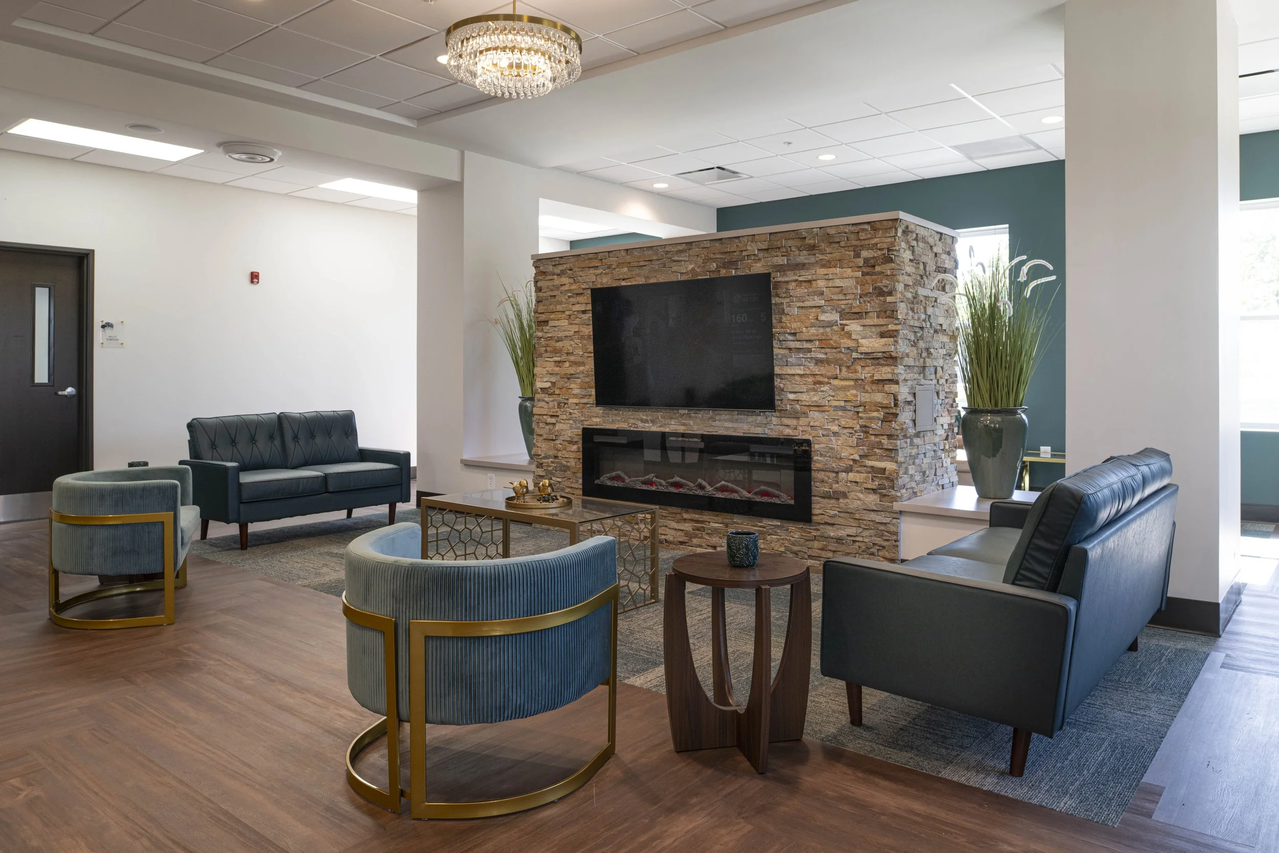 A modern hotel lobby with a stone fireplace, a large flat-screen TV, blue and black leather sofas, striped armchairs with gold accents, a wooden side table, and tall green plants in vases.
