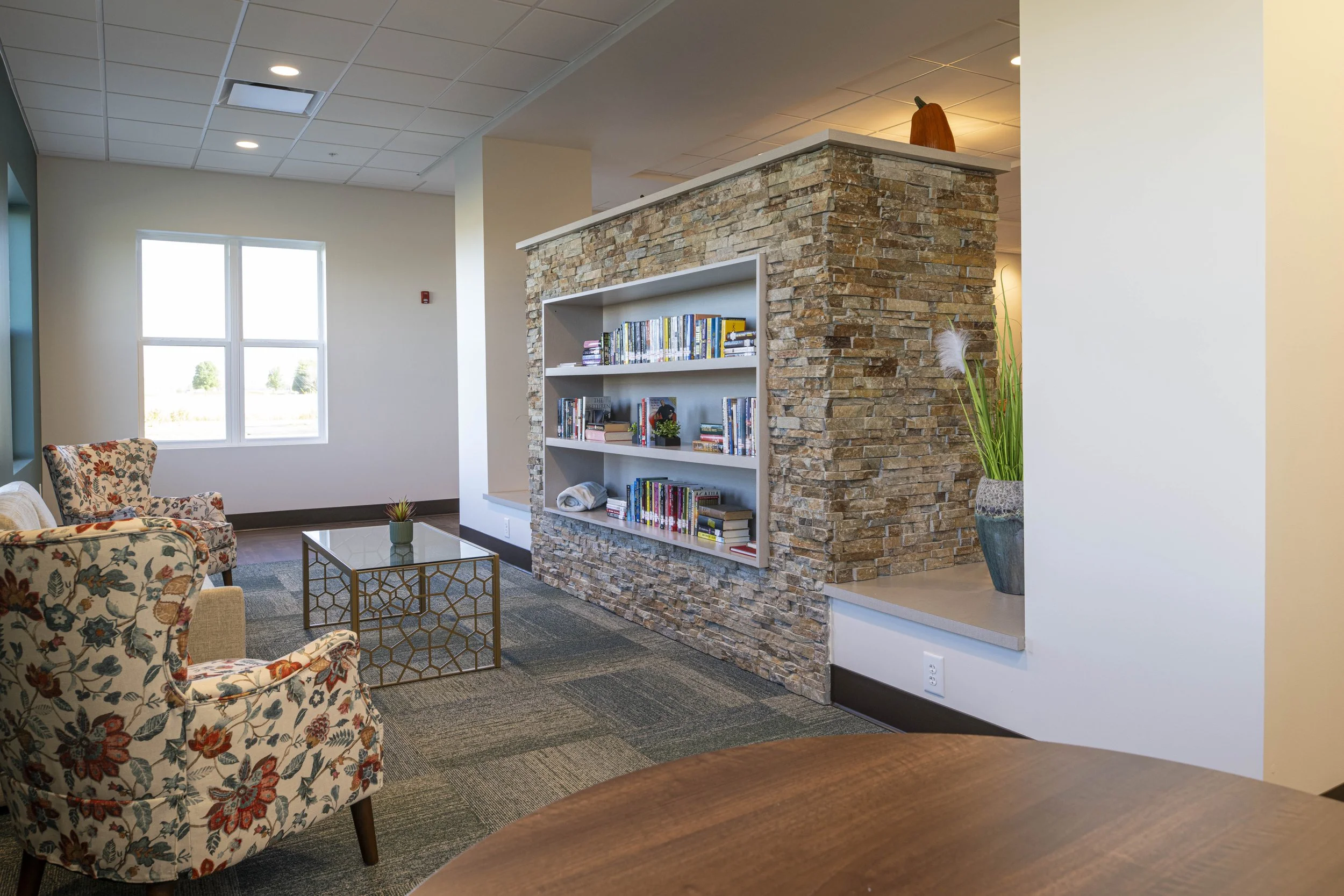 Cozy lounge area with patterned armchairs, a glass coffee table, and a stone wall with built-in bookshelf filled with books. Large window lets in natural light, and there is a tall potted plant on a ledge.