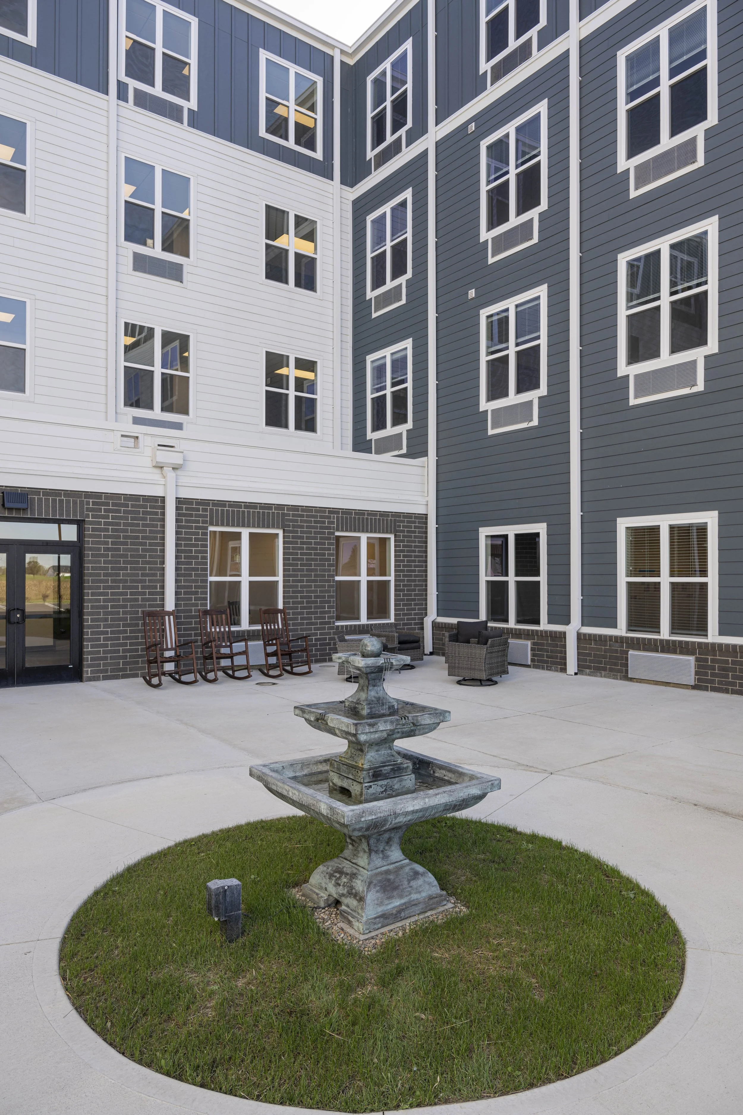 Courtyard with a stone fountain, outdoor seating, and rocking chairs surrounded by a multi-story modern building with blue and white exterior walls and multiple windows.