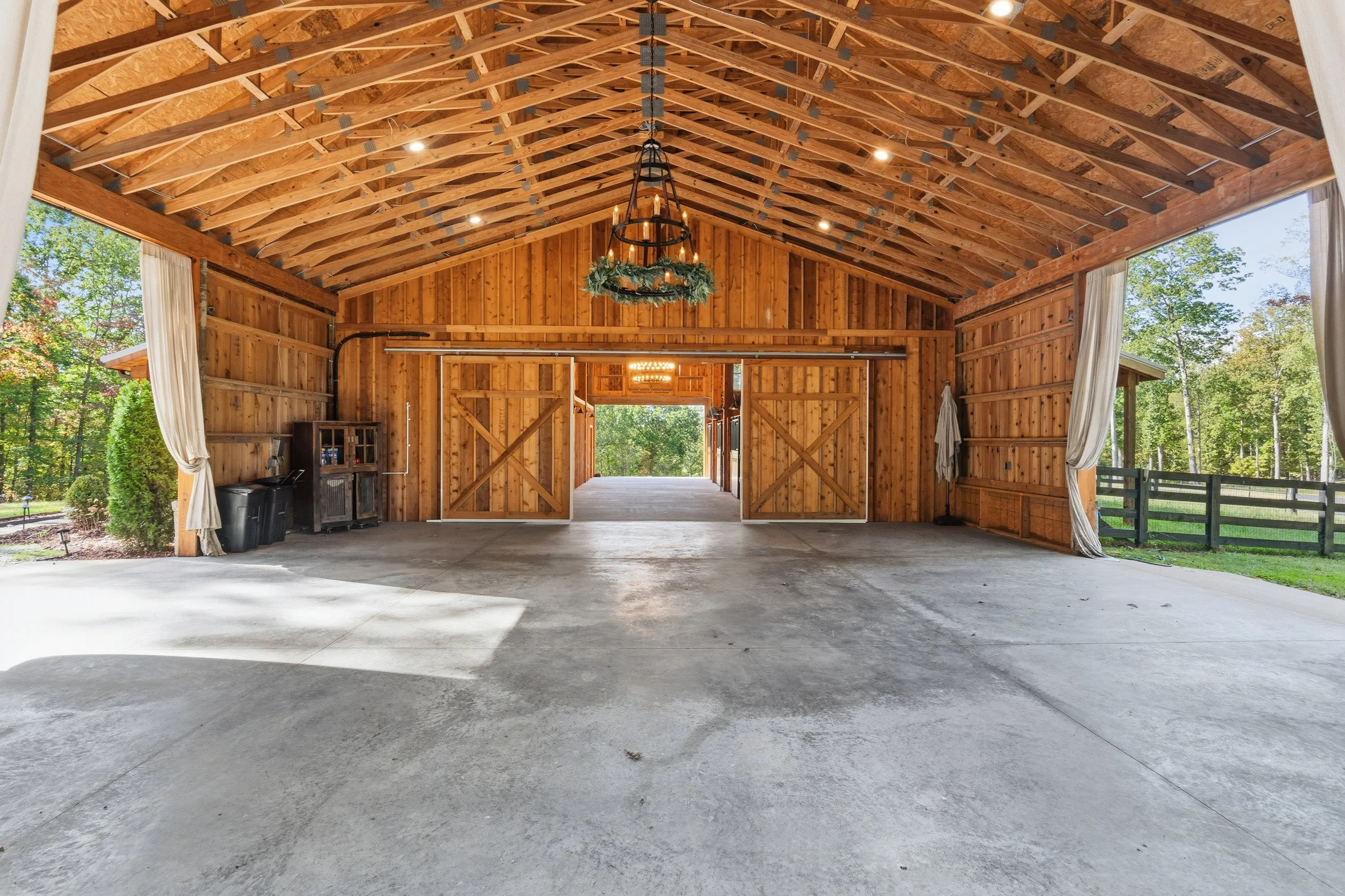 A large wooden barn interior with a high vaulted ceiling, open sliding barn doors revealing a view of green trees outside, concrete floor, and some small furniture and decorations near the sides.