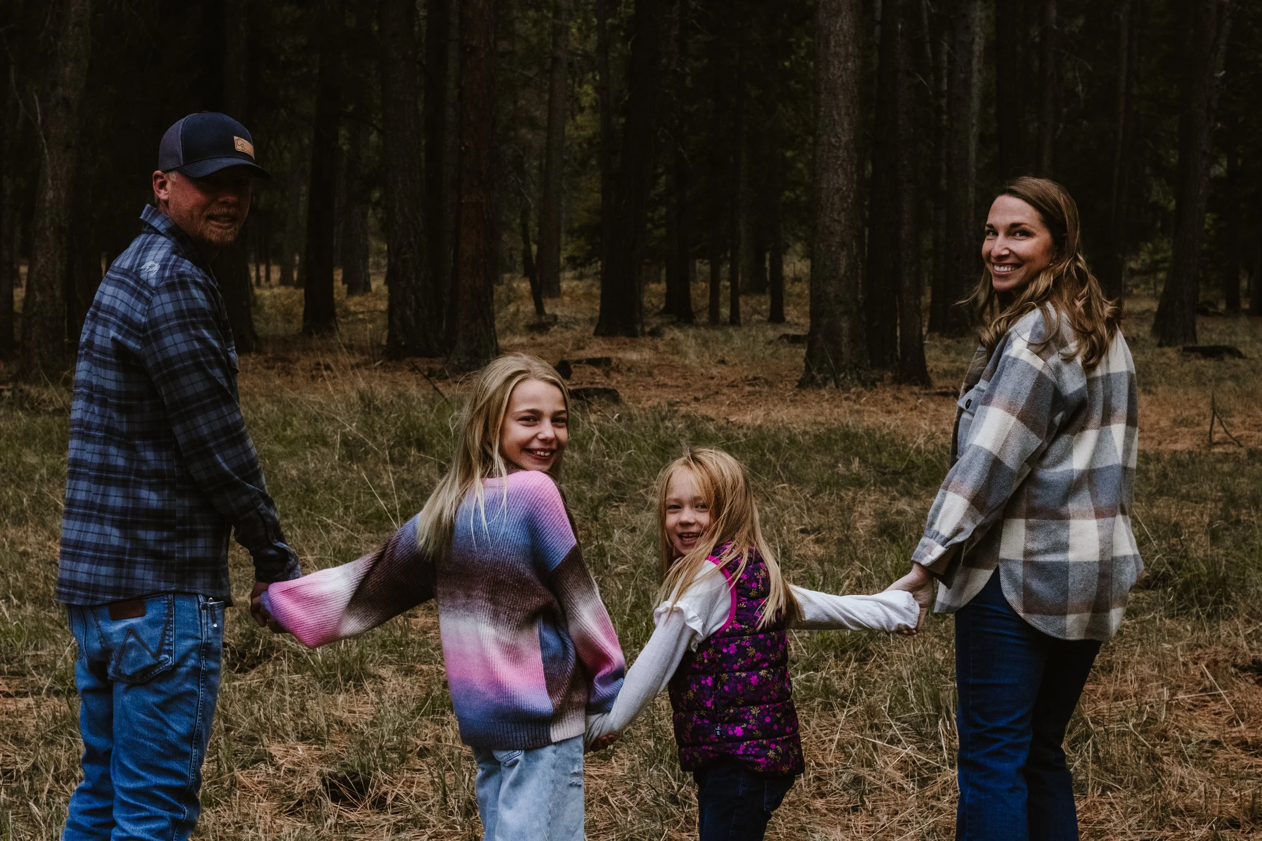 A family of four holding hands in a forest, smiling and enjoying outdoor activity.