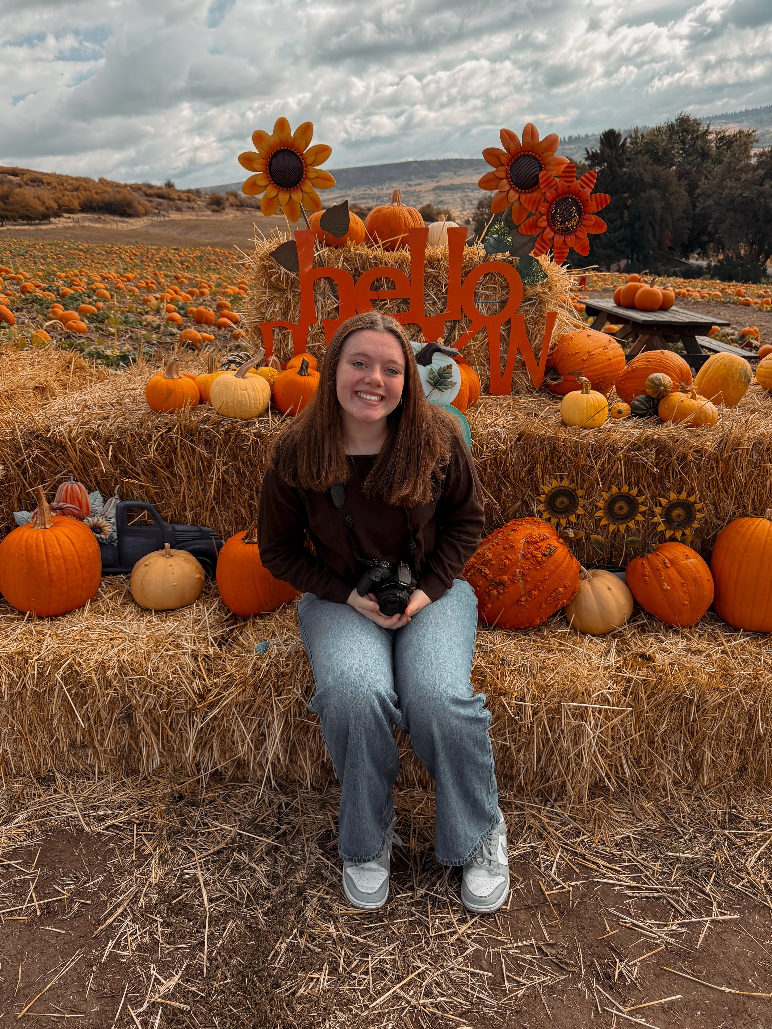 A young woman with brown hair, wearing a dark jacket, sitting on hay bales at a pumpkin patch. She is smiling and holding a camera. Behind her is a large fall display with pumpkins, sunflowers, and a sign that says "hello". The pumpkin patch extends into the background under a cloudy sky.