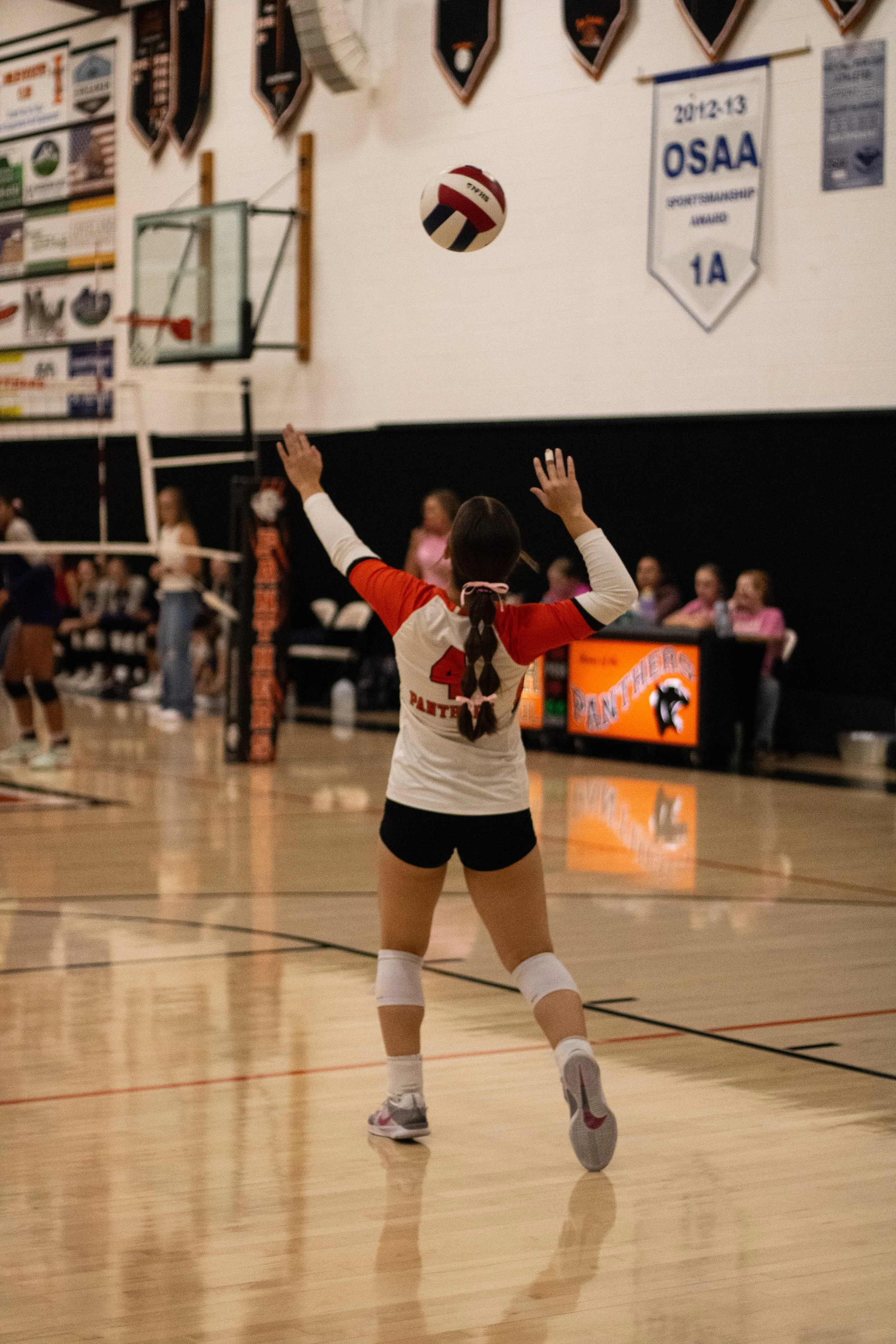 A girl in a volleyball uniform, with jersey number 4, preparing to serve a volleyball in a gymnasium during a match, with spectators and teammates in the background.