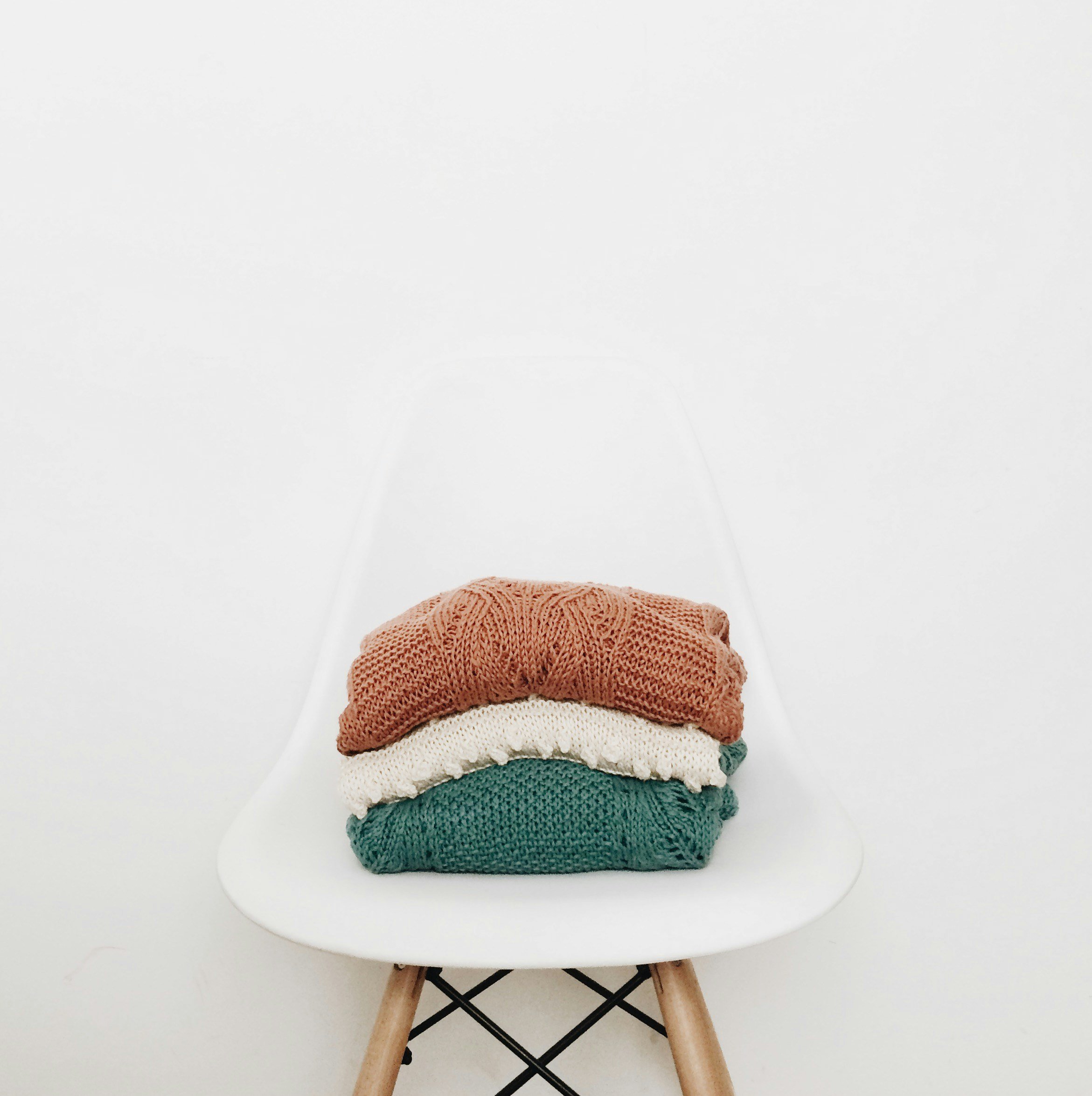 Stack of three knitted sweaters in brown, white, and green on a white chair with wooden legs against a white background.