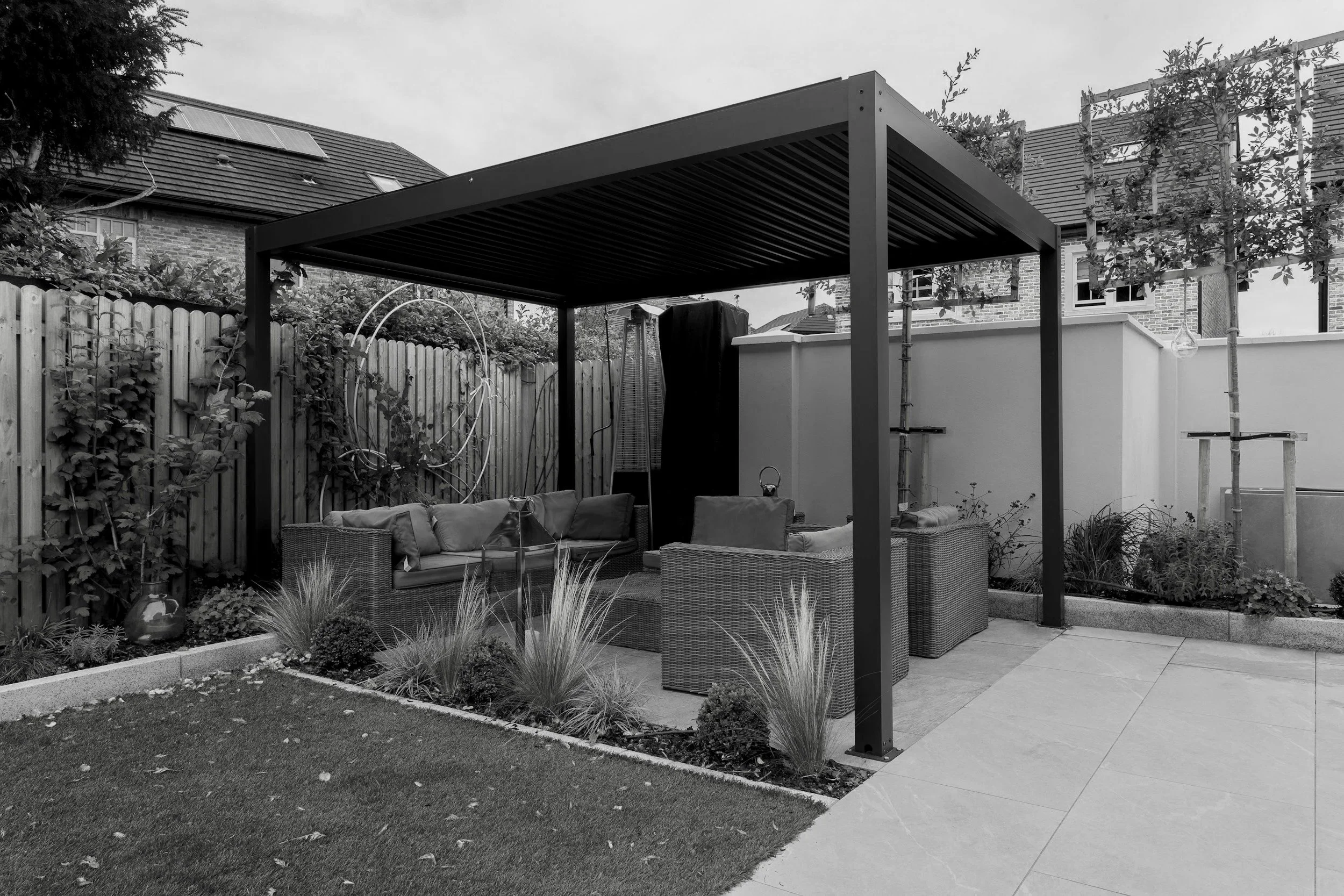 Backyard patio area with outdoor furniture under a pergola, plants along the fence, and neighboring houses in the background.
