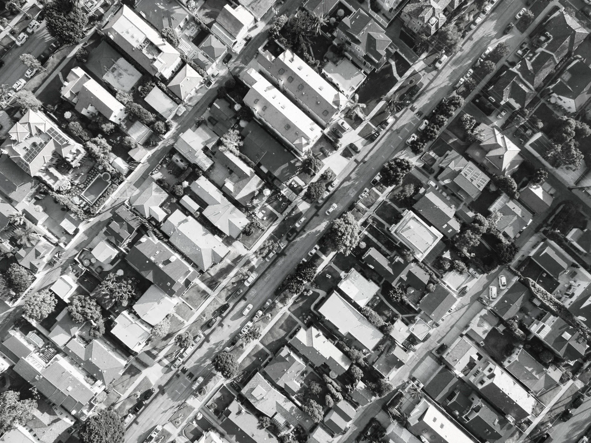 An aerial view of a residential neighborhood with houses, trees, streets, and parked cars in black and white.