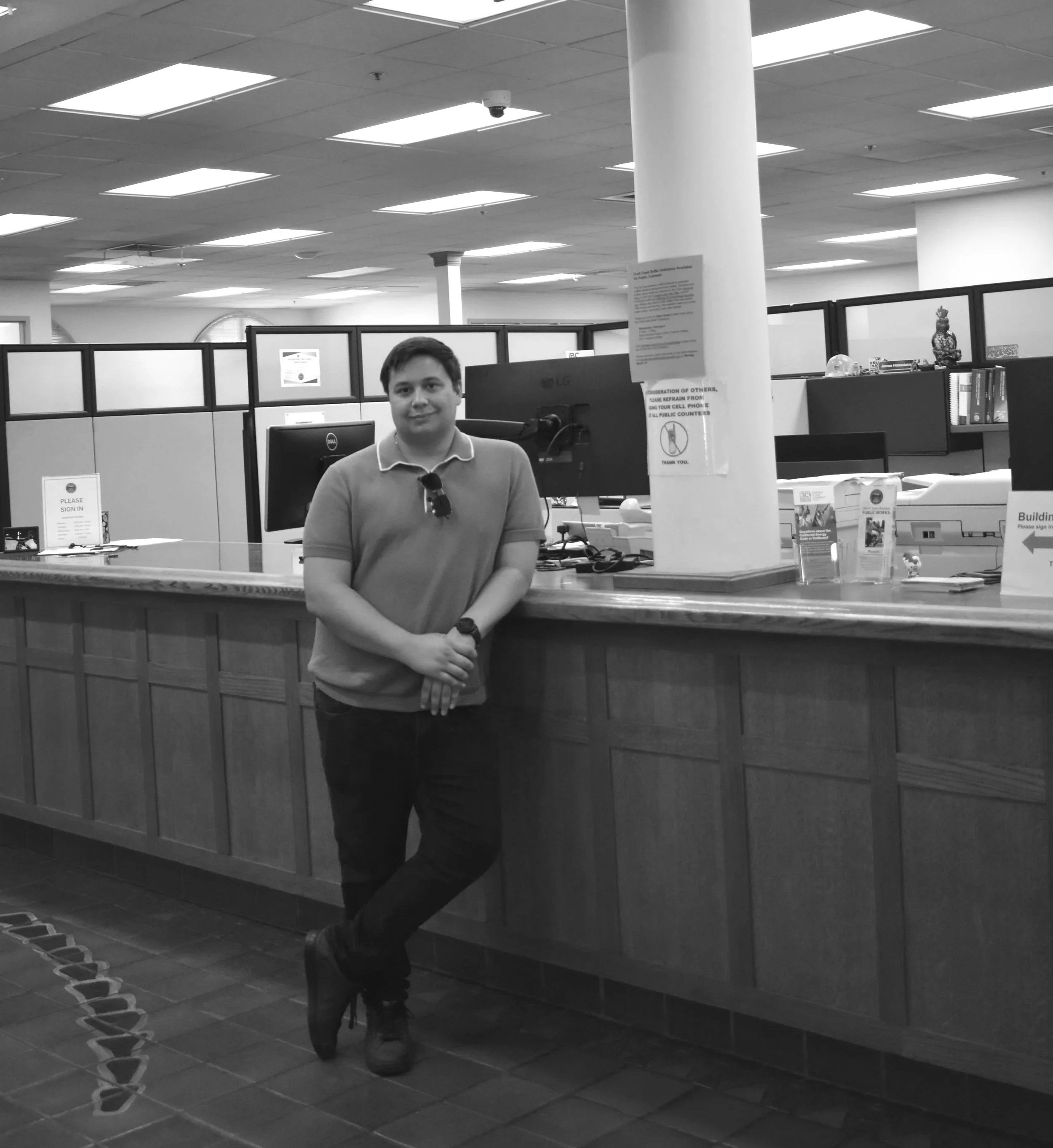 A young man standing at a reception desk in an office or government building, with a computer monitor behind him and cubicle partitions in the background.