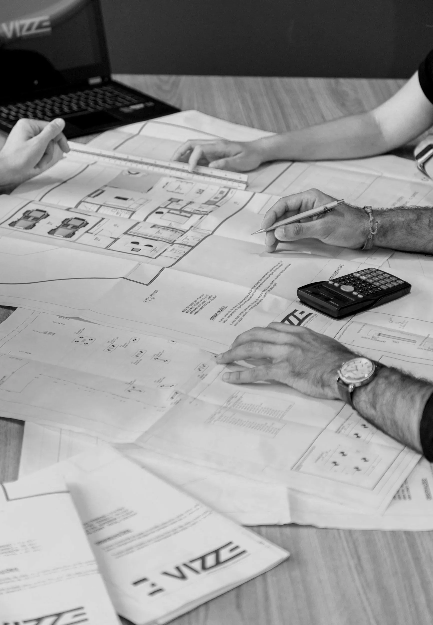 People working on architectural plans and blueprints spread out on a table, with a calculator, pen, ruler, and laptop present.