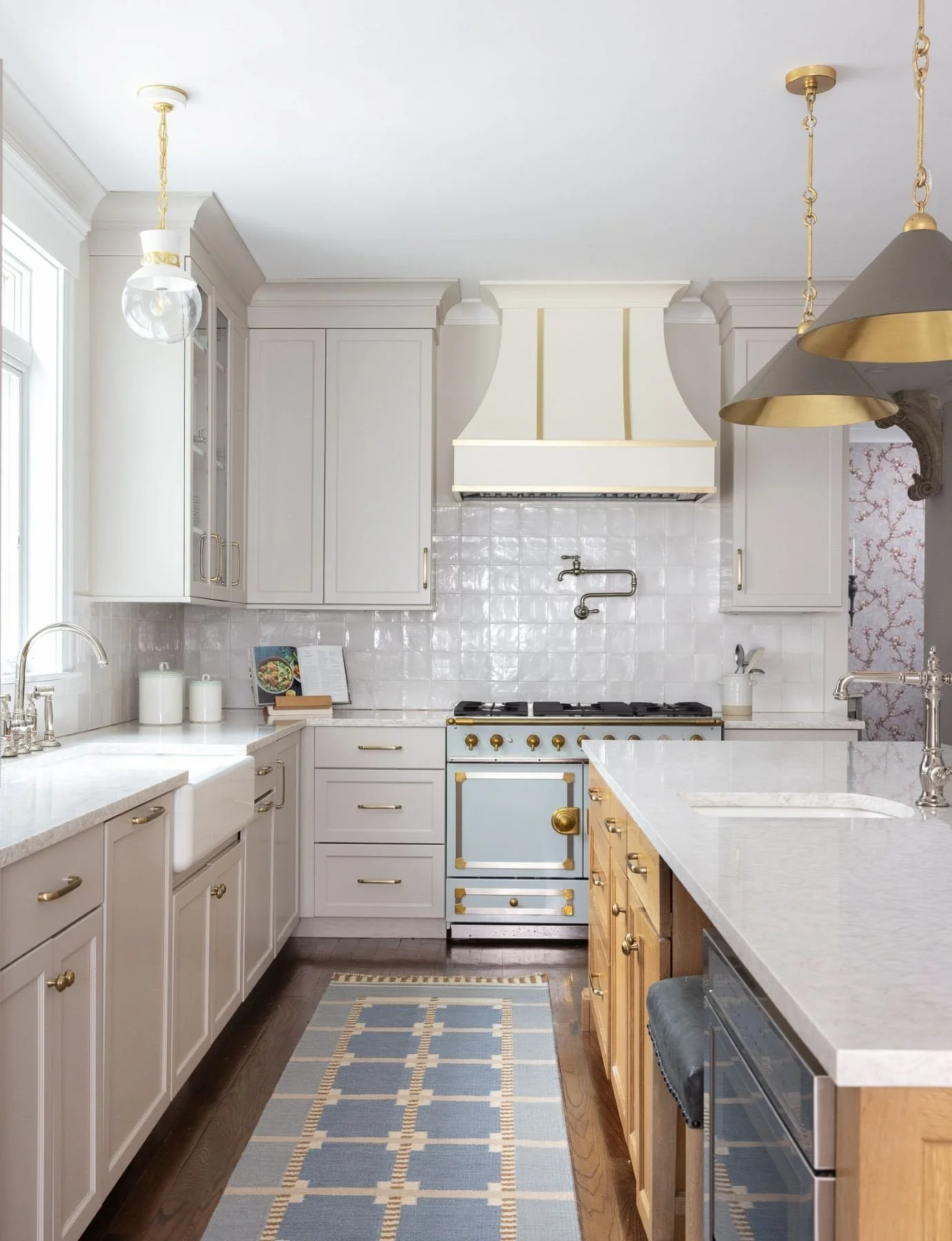 A kitchen with white cabinets with gold handles, a white range hood with gold accents, a vintage blue oven, a white countertop with a kitchen sink, a blue and beige patterned rug, and gold pendant lights.