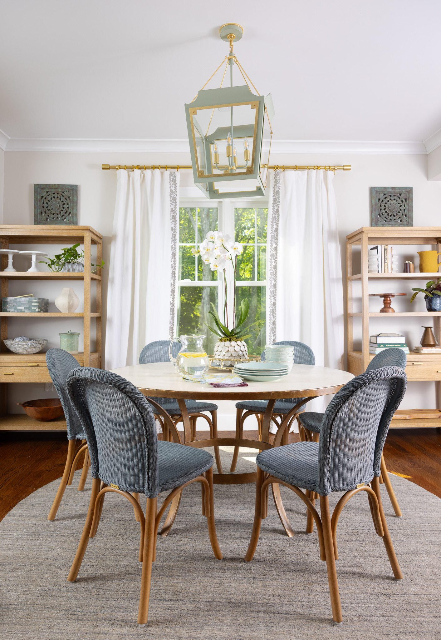 A dining room with a round wooden table, six chairs with gray upholstery, white curtains, wooden bookshelves, a modern chandelier, and a window with greenery outside.