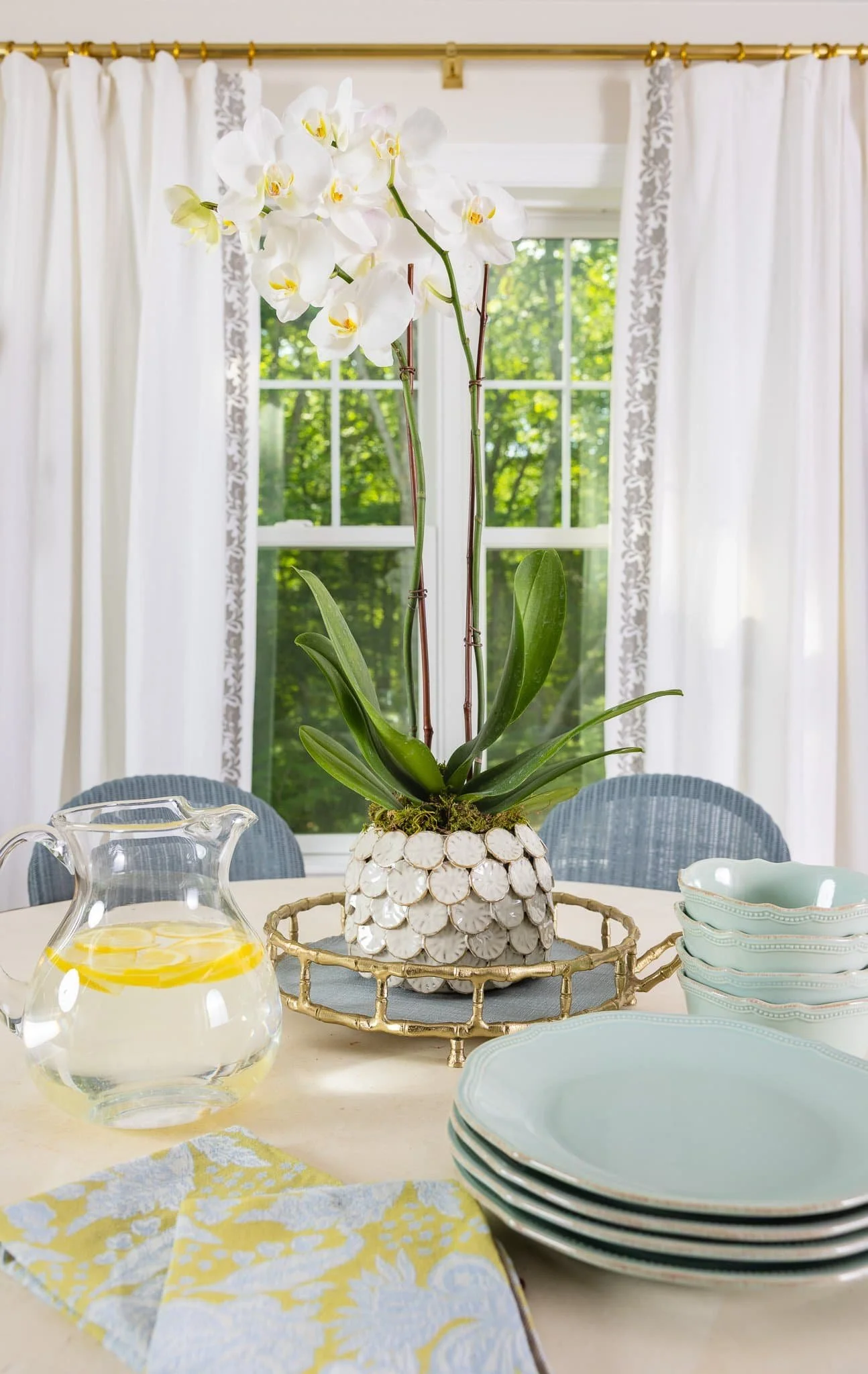A white orchid plant in a decorative pot on a dining table, with stacked plates and a pitcher of lemonade nearby, in front of a window with white curtains.