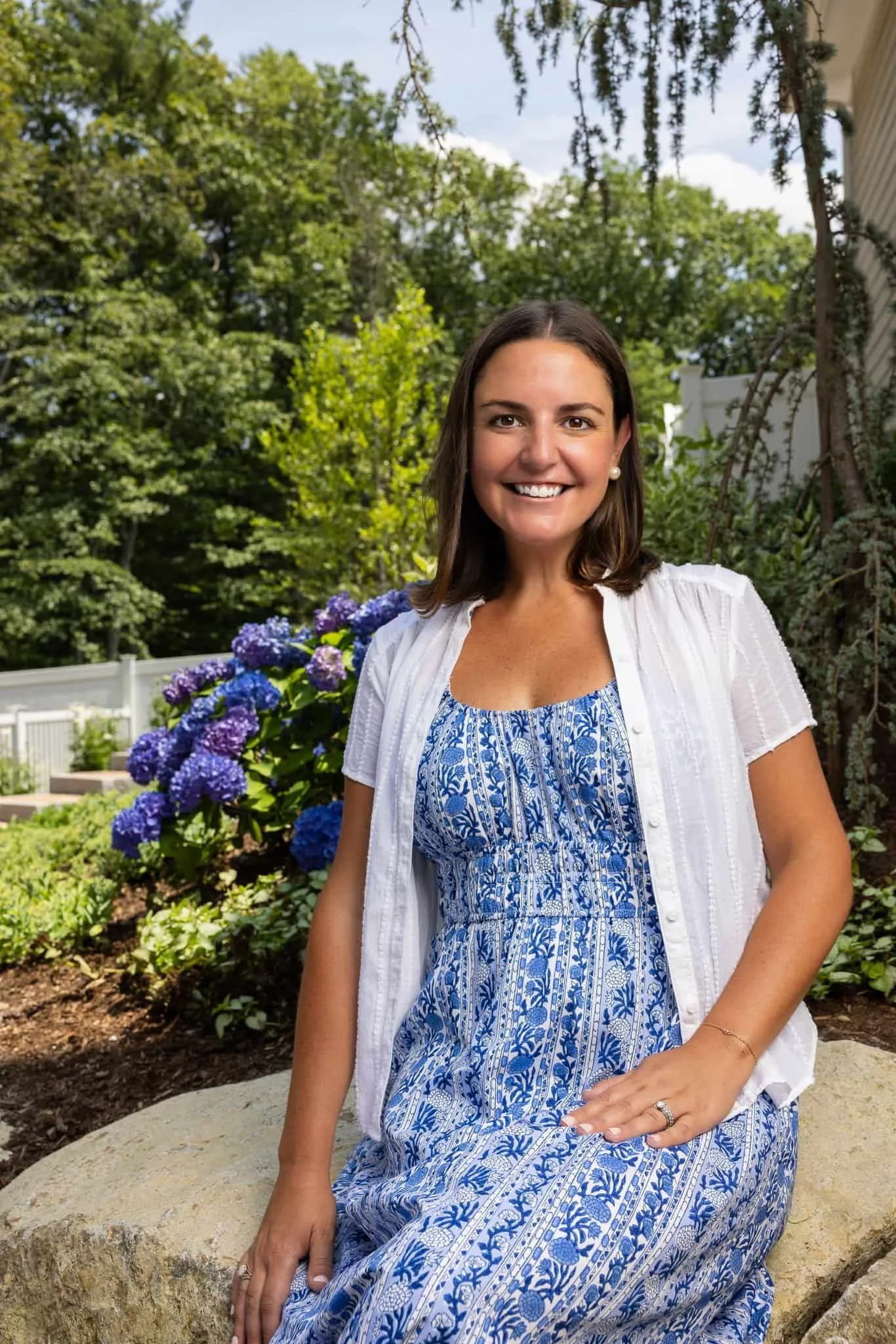 A woman in a blue and white floral dress and white shirt sitting on a stone in a garden with purple hydrangea flowers and green trees in the background, smiling at the camera.