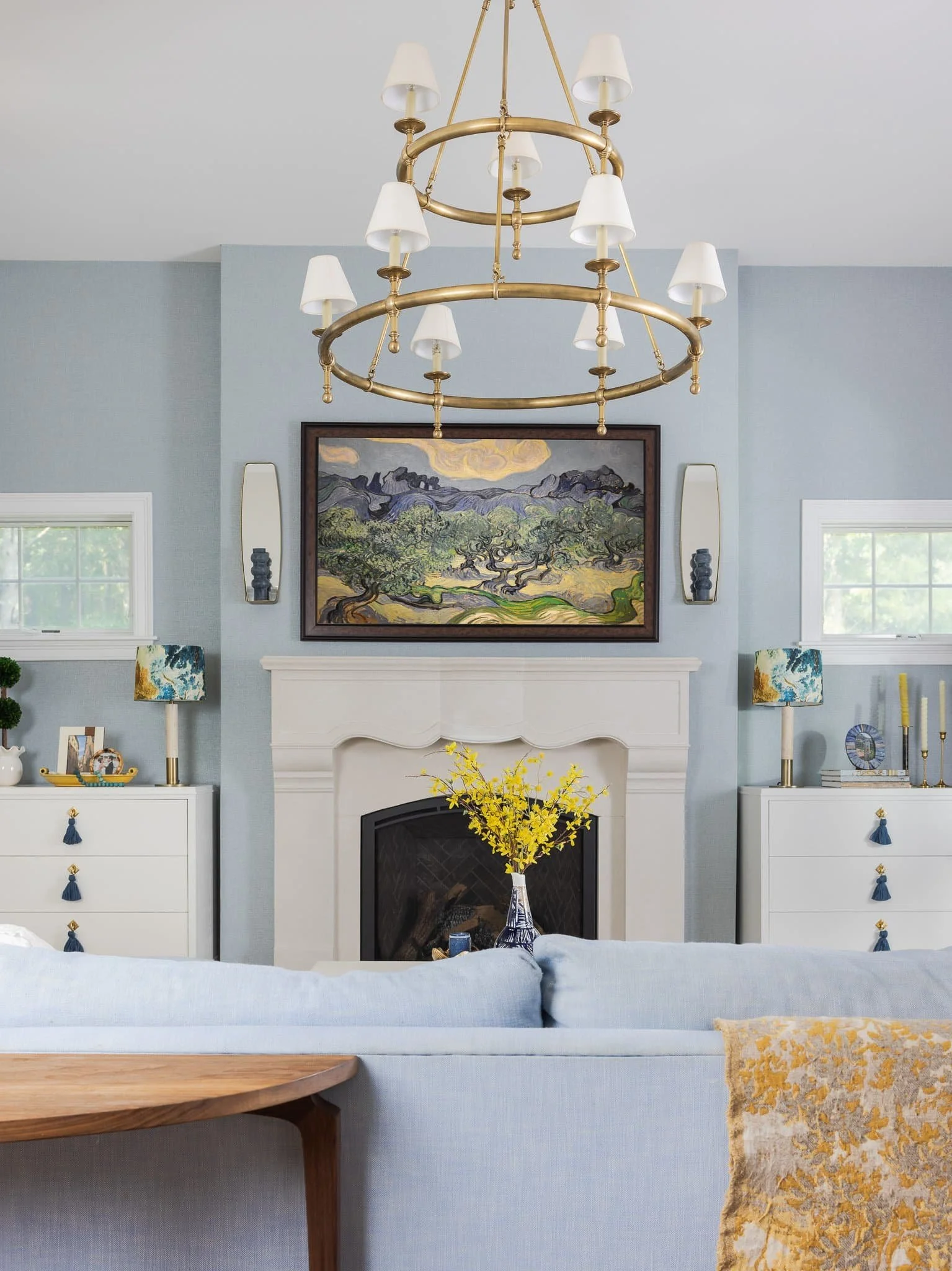 Living room with a white fireplace, a chandelier, two white dressers, colorful lamps, and a painting of a landscape above the fireplace.