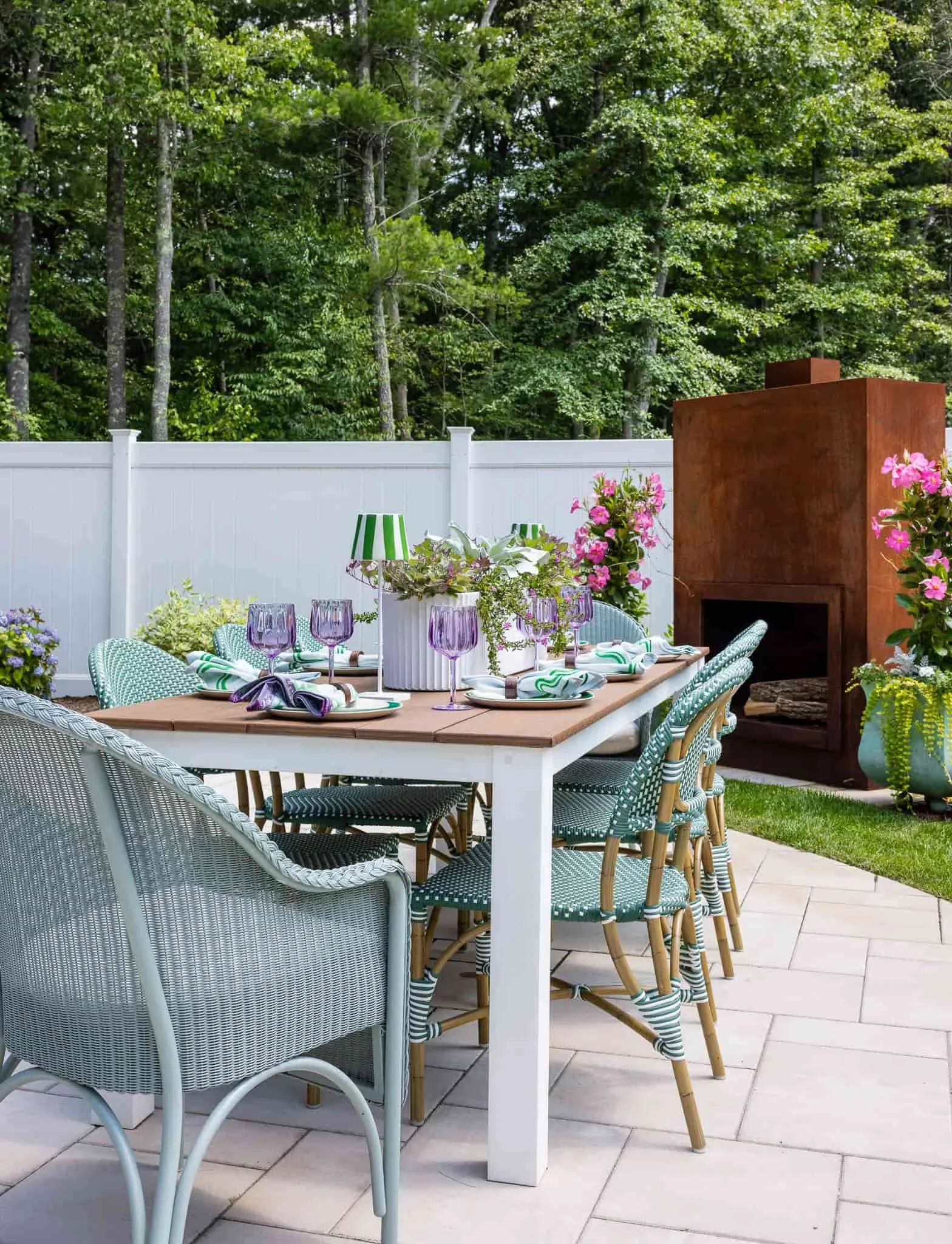 An outdoor patio table set for a meal with purple wine glasses, green and white striped napkins, and white plates, surrounded by wicker and rattan chairs, with a fire pit and potted flowers, against a white fence and lush green trees.