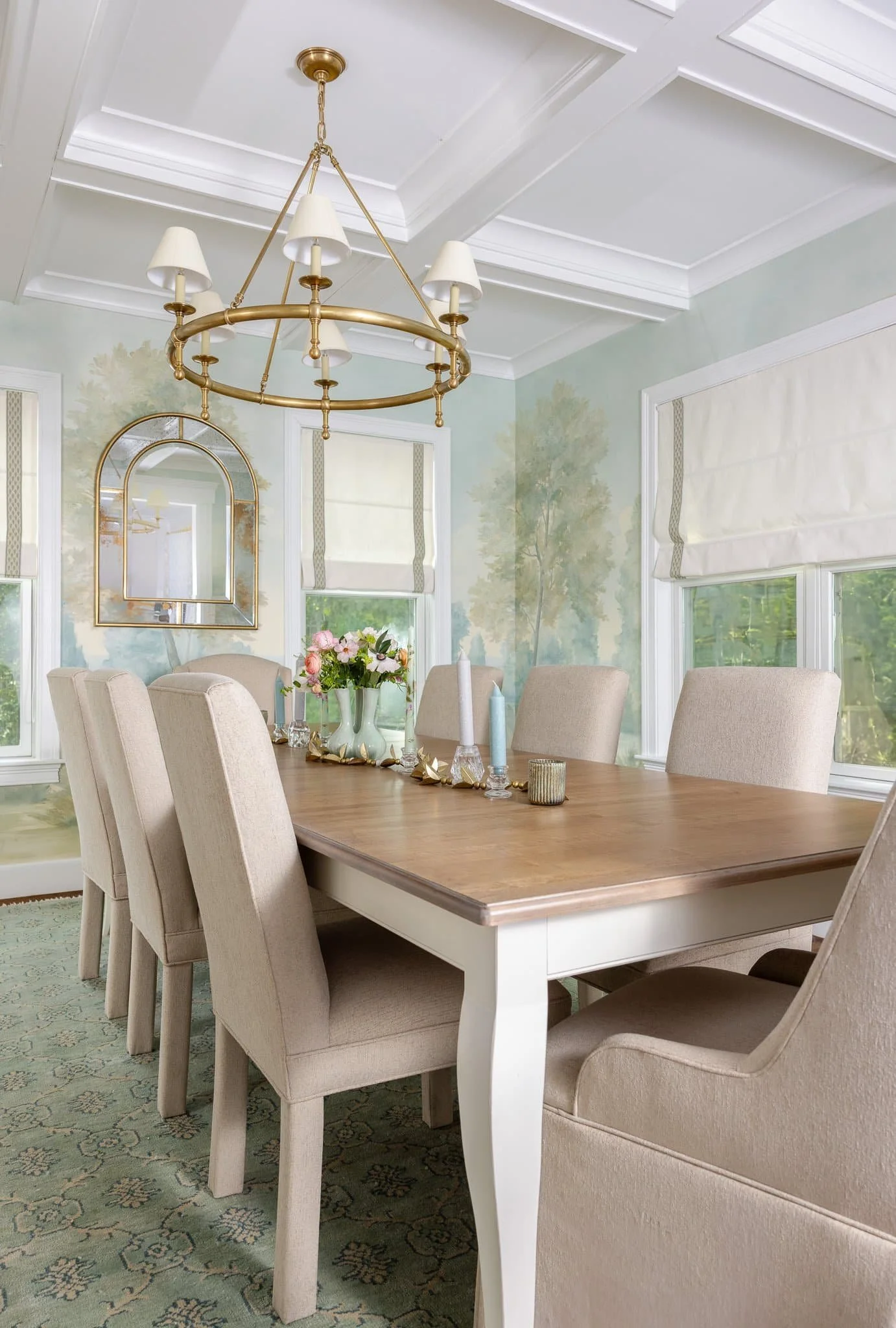 A dining room with a wooden table, beige upholstered chairs, a chandelier with white shades, a wall mirror, a painted landscape mural, and windows with Roman shades.