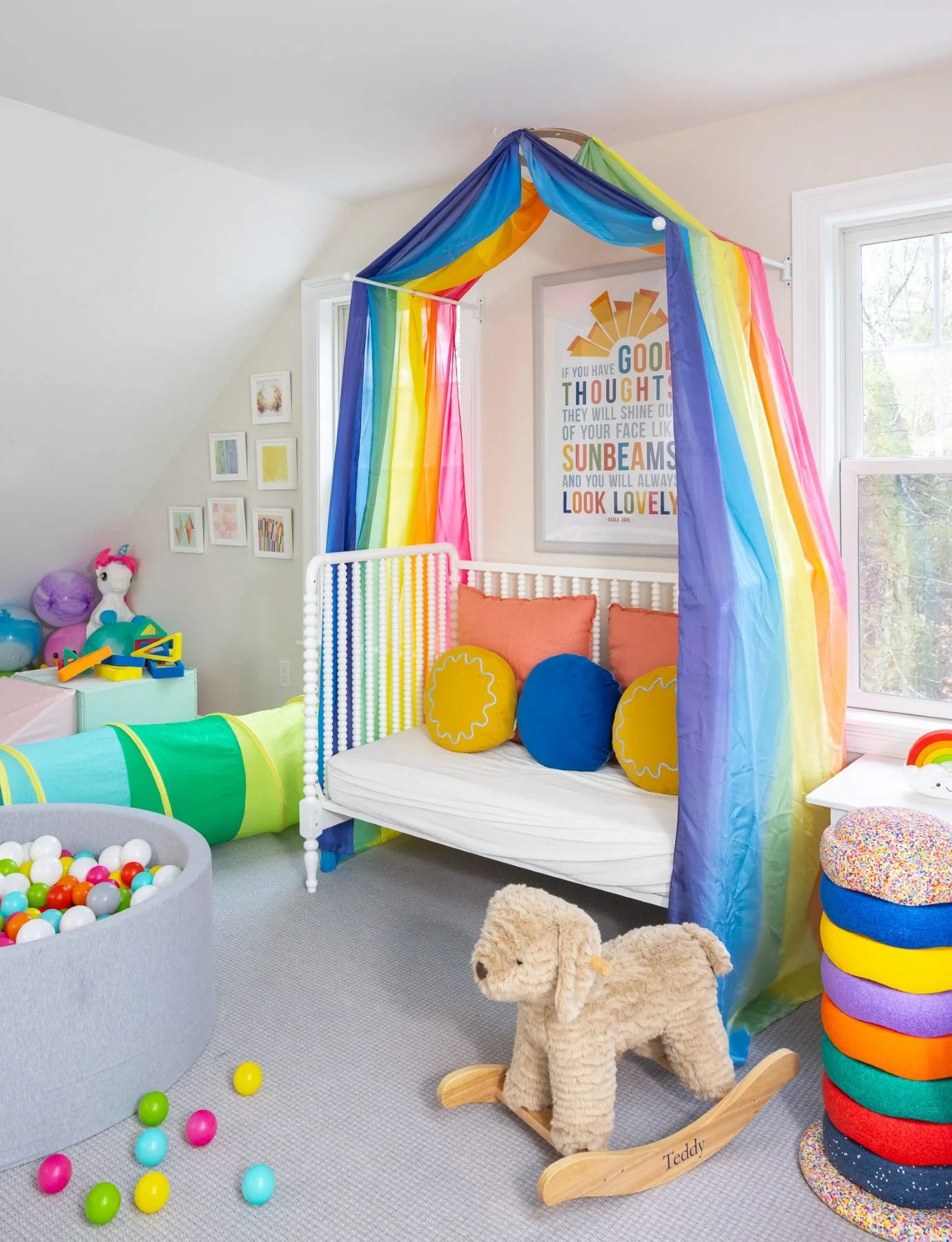 Colorful children's playroom with rainbow canopy over a crib, stuffed animals, toy balls, and cheerful decor.